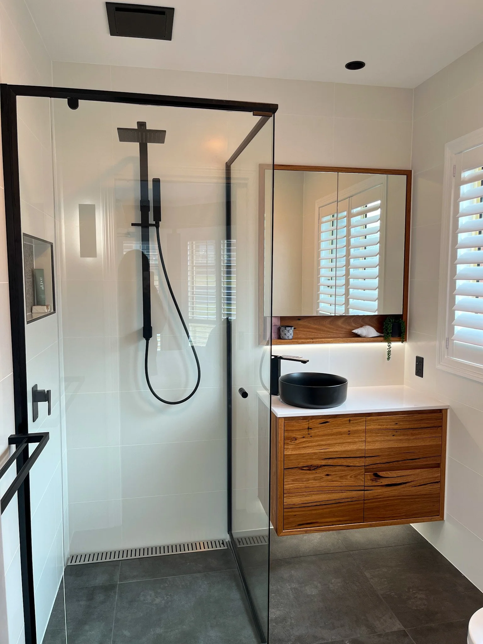 Modern bathroom with a walk-in shower, black fixtures, wooden vanity with a black vessel sink, mirror, and white plantation shutters on windows.