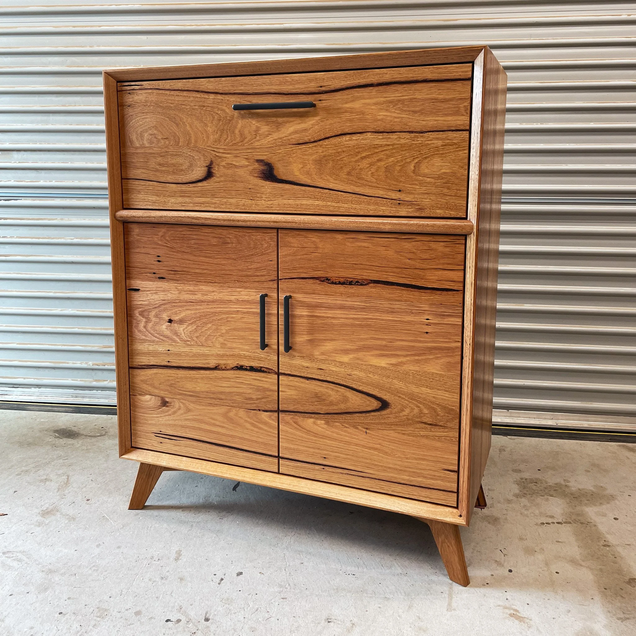 A mid-century modern wooden cabinet with four drawers, standing on angled legs, is set against a corrugated metal wall in an industrial space.