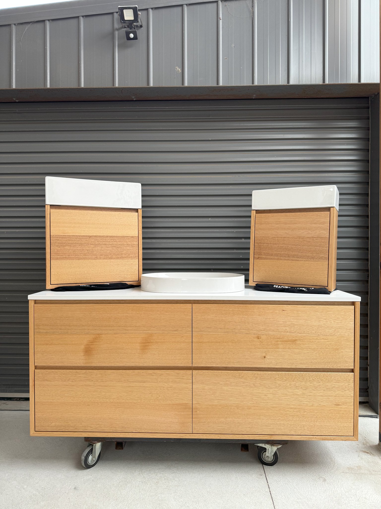 A mobile wooden sideboard on wheels with two white shelves and a white circular tray on top, set against a gray corrugated metal wall.
