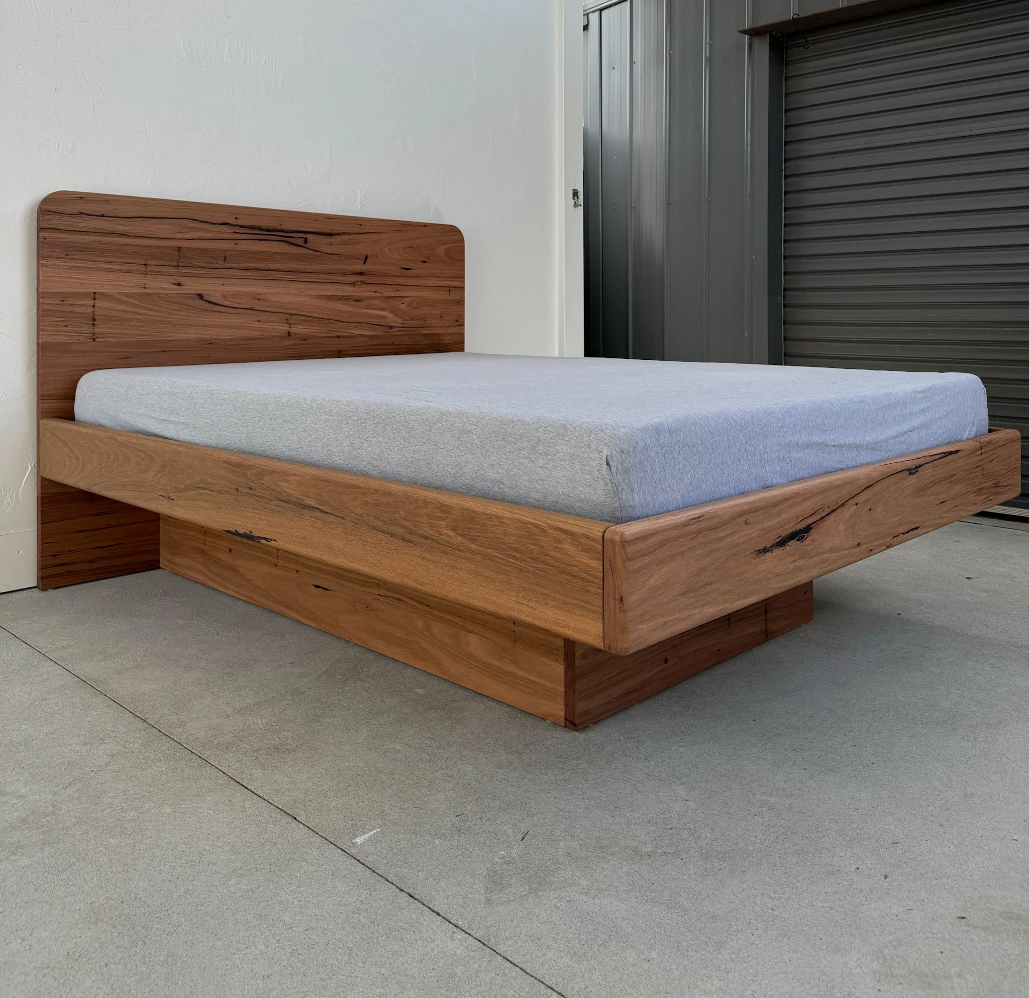 A timber bed frame, ifs floating, recycled hardwood, with a light-colored mattress, positioned next to a white wall and a metallic gray garage door.