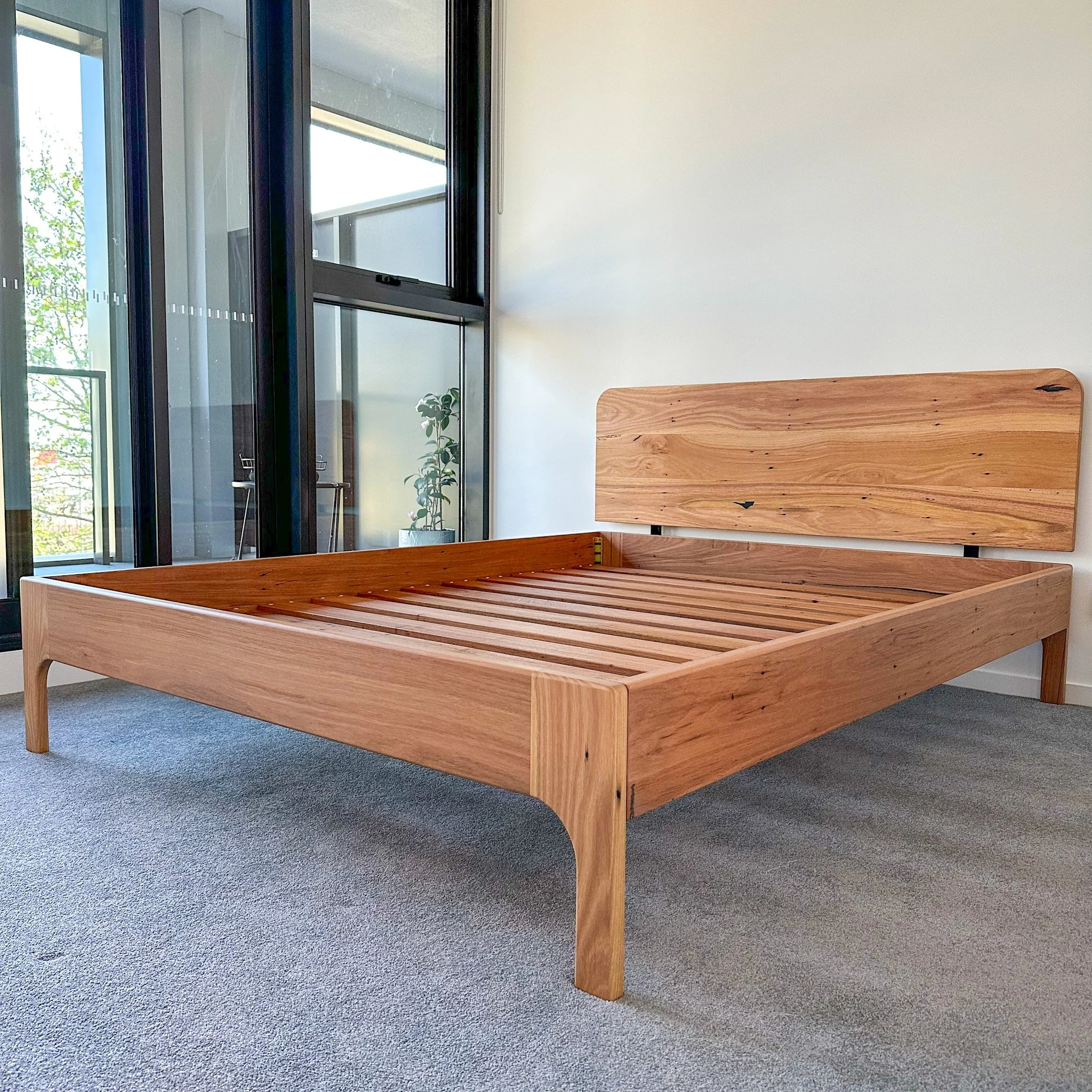 Unfinished wooden bed frame in a room with large windows, white walls, and a gray carpet.