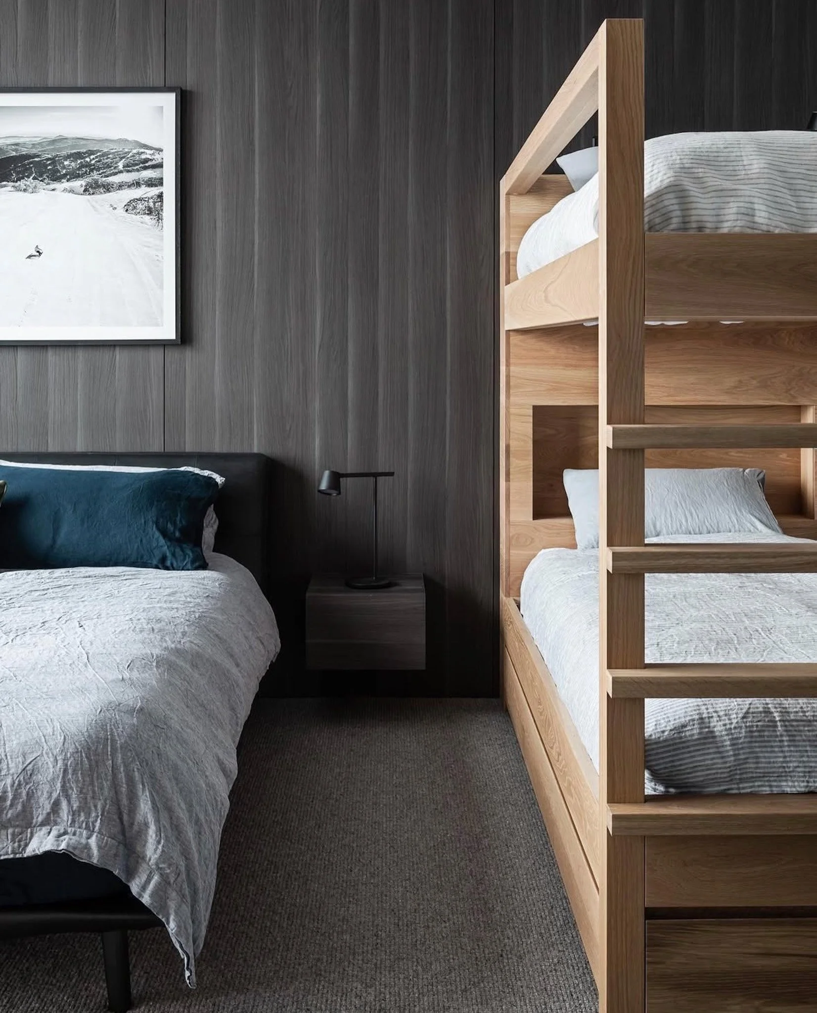 A bedroom with a bed, a nightstand, a black lamp, dark wood wall, framed black and white landscape photo, and a wooden bunk bed