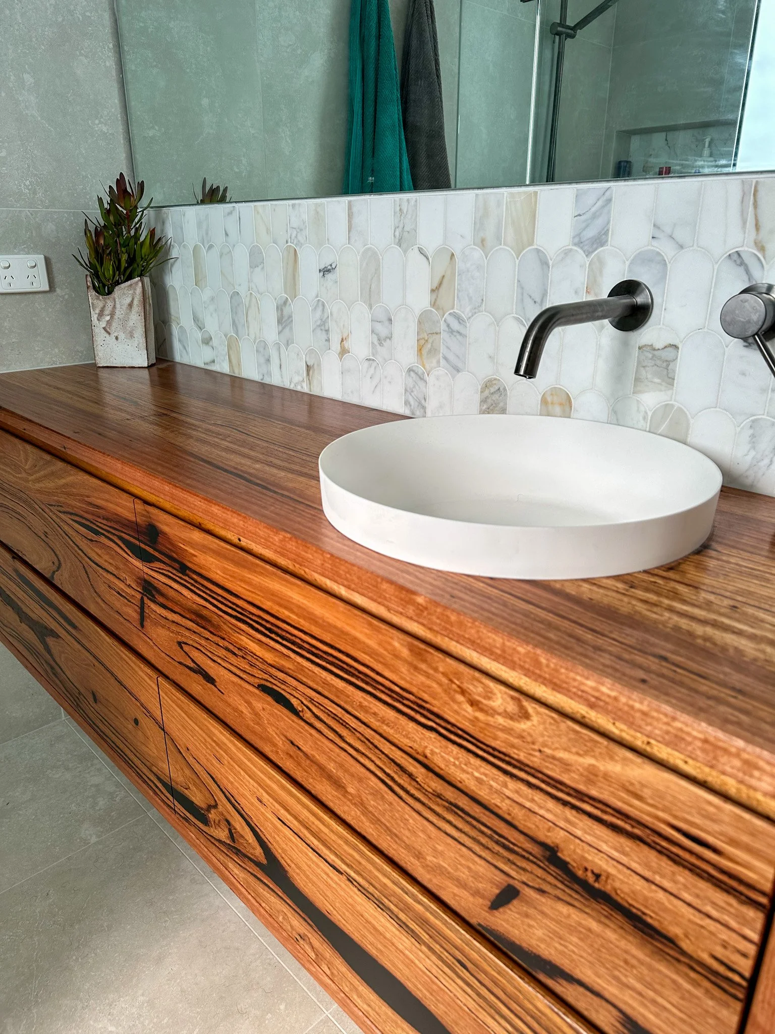 A bathroom vanity with a wood grain finish, a white oval sink, and a black wall-mounted faucet. A marble tile backsplash with a pattern of elongated, scalloped shapes runs above the vanity. There is a potted plant with dark green and reddish leaves o