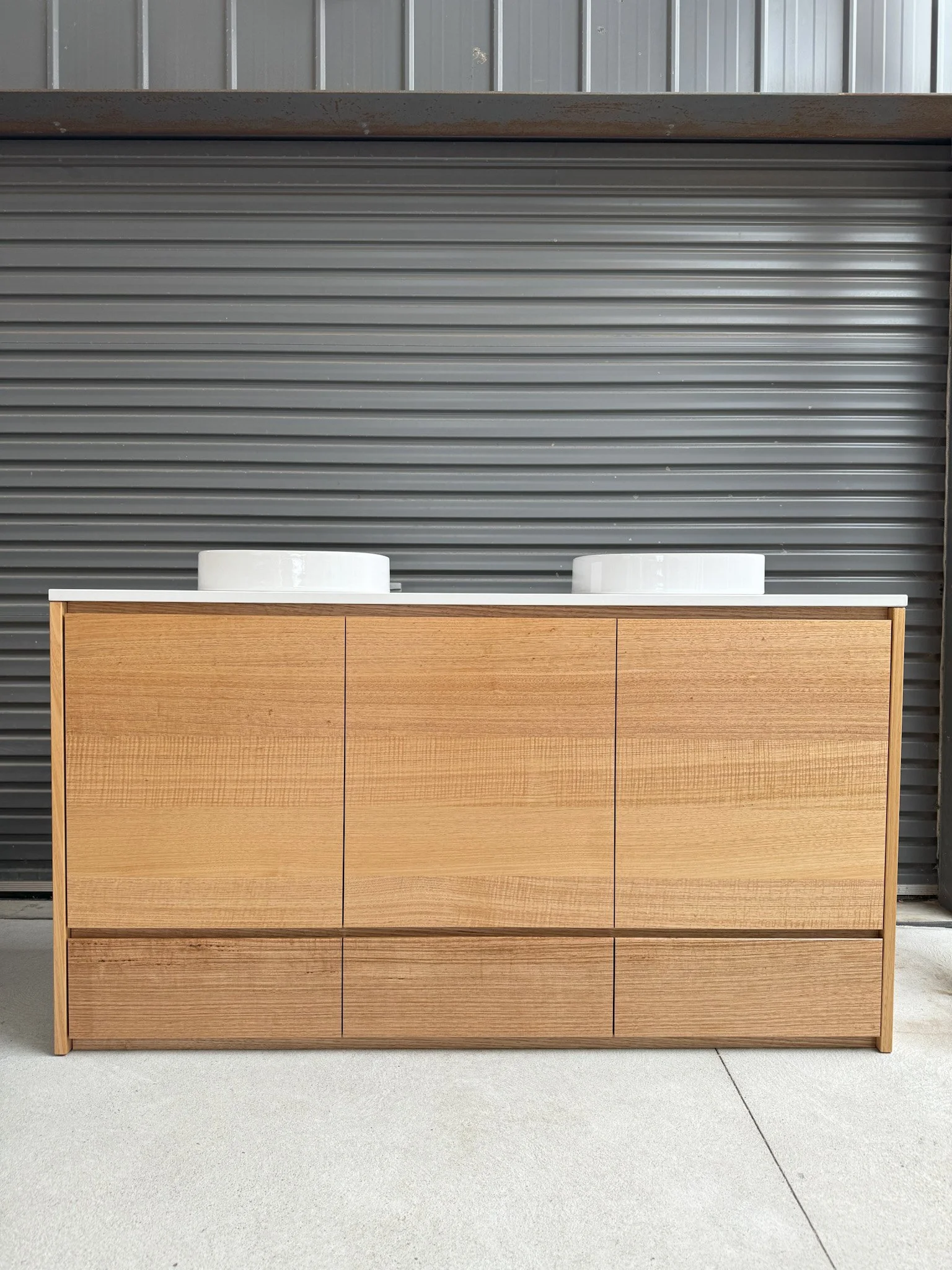 Timber vanity with six drawers and two white bowls on top, set against a gray metal wall.