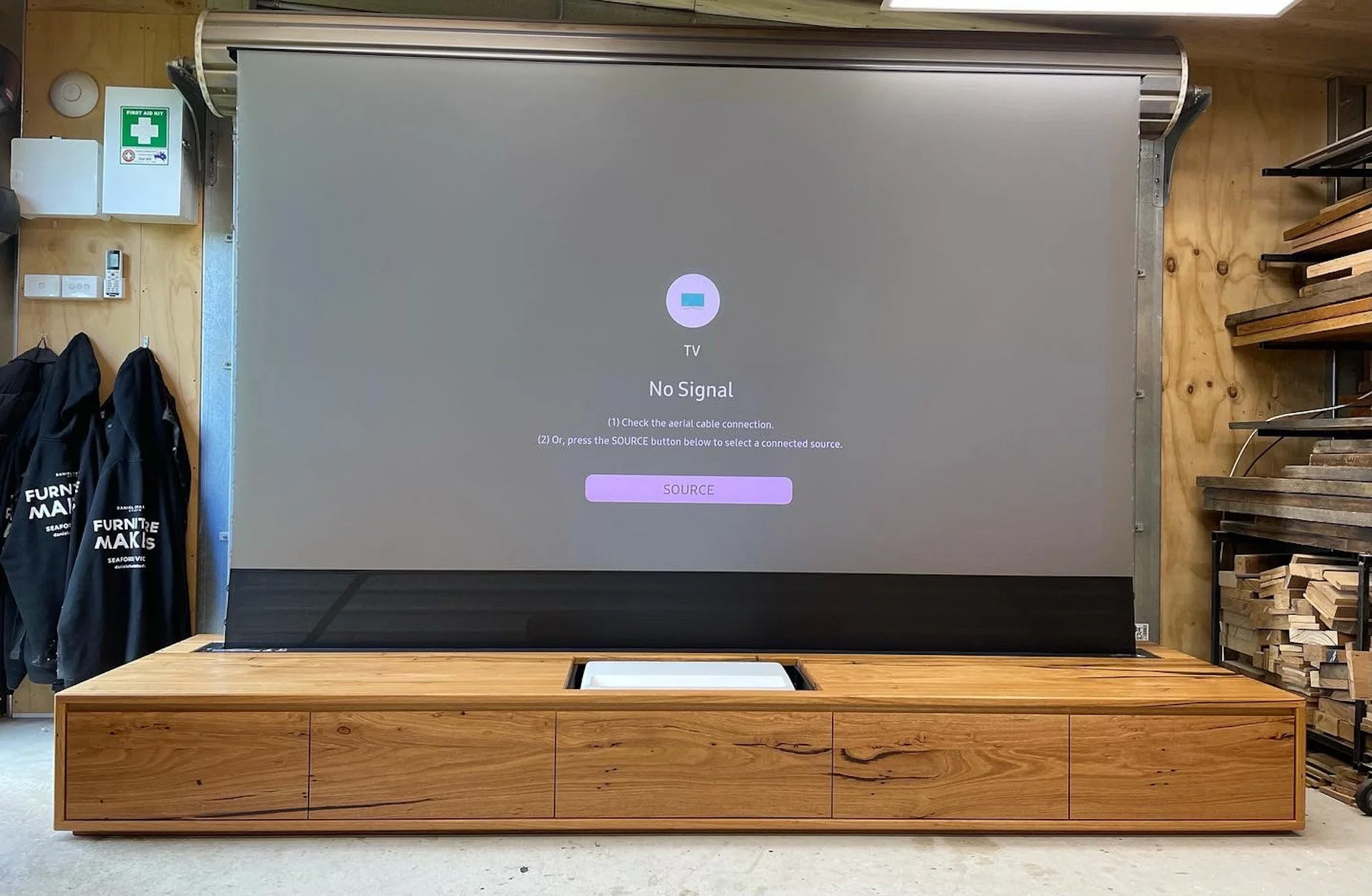A large flat-screen TV mounted on a wooden table with a message reading 'No Signal' and instructions to check the aerial cable connection or press the source button. To the left, there are black jackets hanging and some first aid supplies on the wall