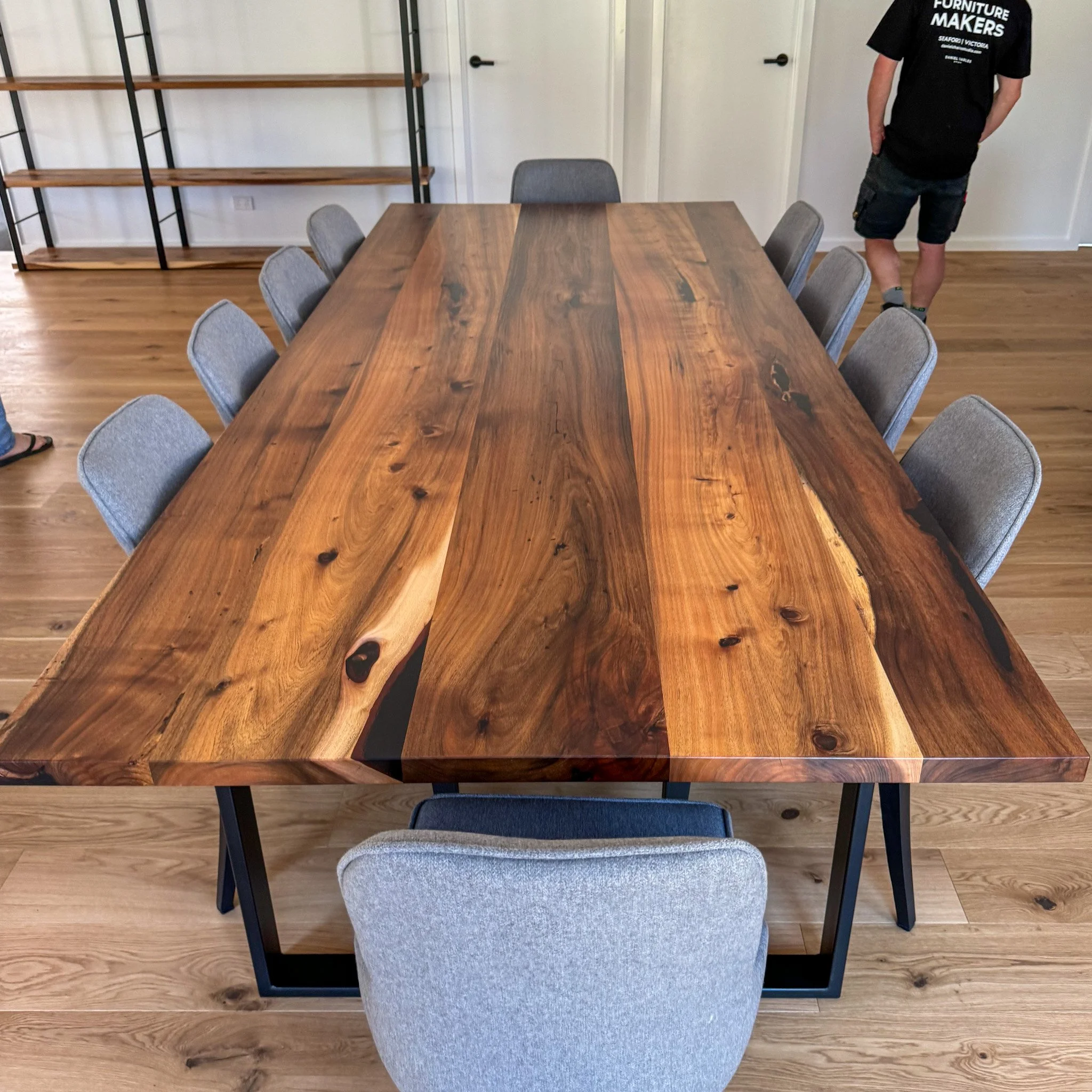 A wooden conference table surrounded by gray upholstered chairs in a room with wood flooring and a shelving unit against the wall. A person in a black t-shirt and shorts is partially visible standing near the door.