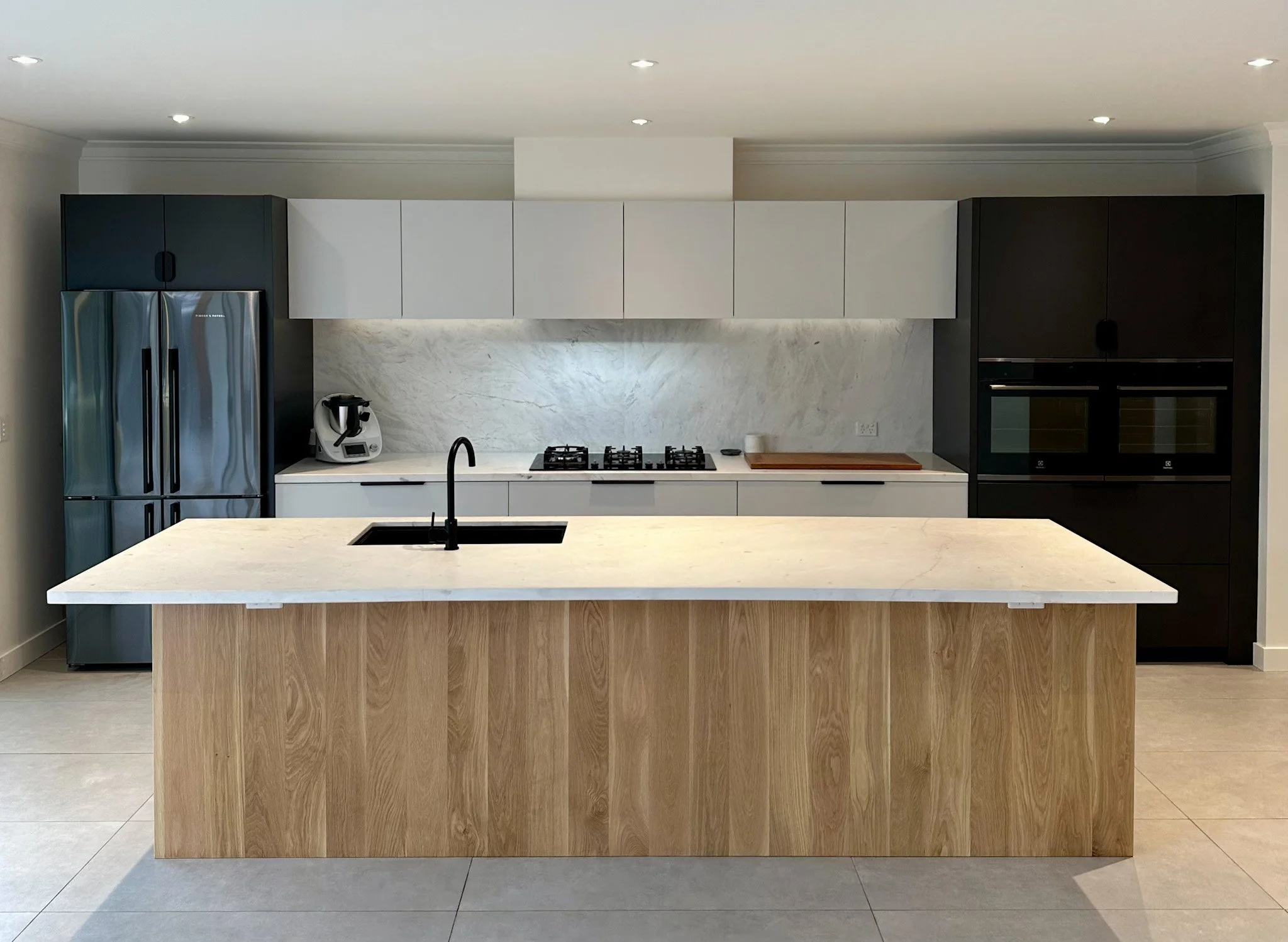 Modern kitchen with a large island, stainless steel refrigerator, white upper and black lower cabinets, marble backsplash, and built-in oven.