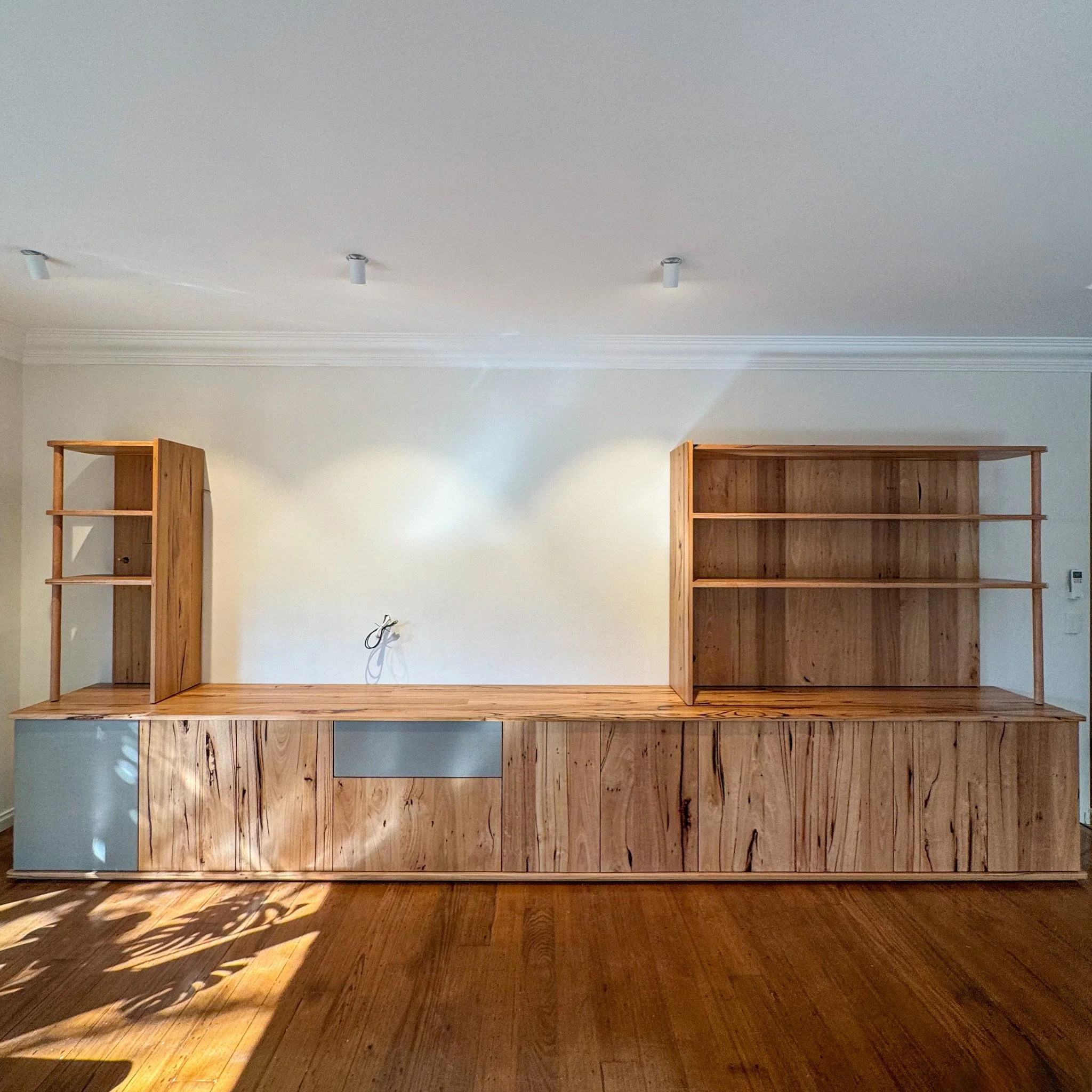 Empty wooden shelves on a beige wall with a shadow of a plant on a wooden floor.