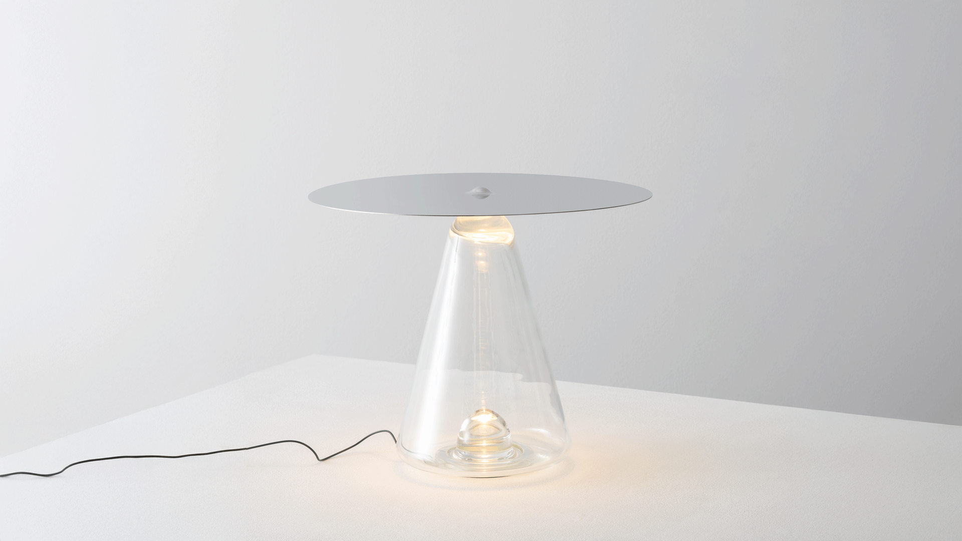 Lampada moderna con base di vetro trasparente e lampadina visibile, luce soffusa, su superficie bianca.