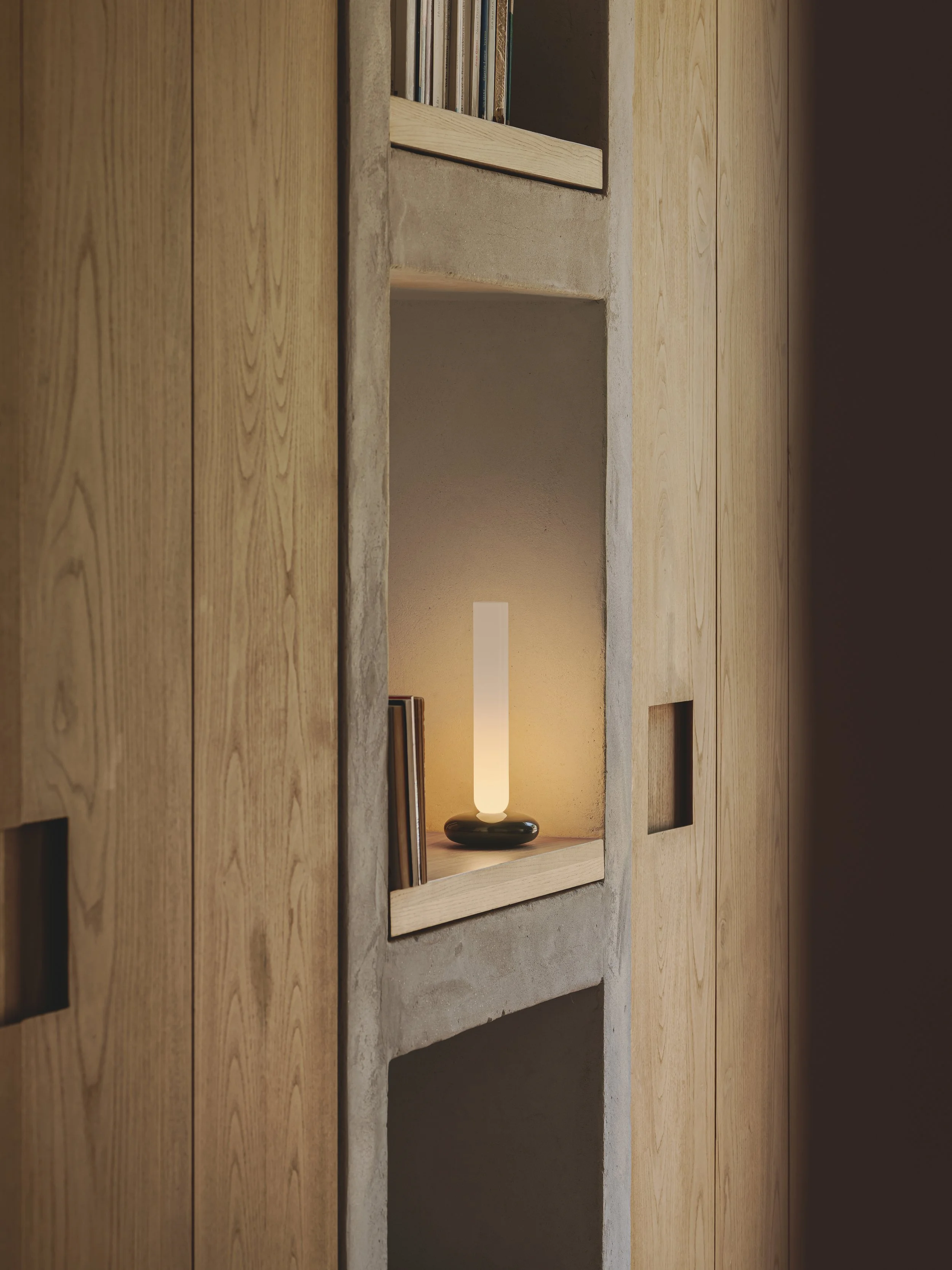 Uno scaffale incassato in una parete, decorato con libri e una lampada a forma di cilindro acceso, con pareti di cartongesso e pannelli di legno attorno.