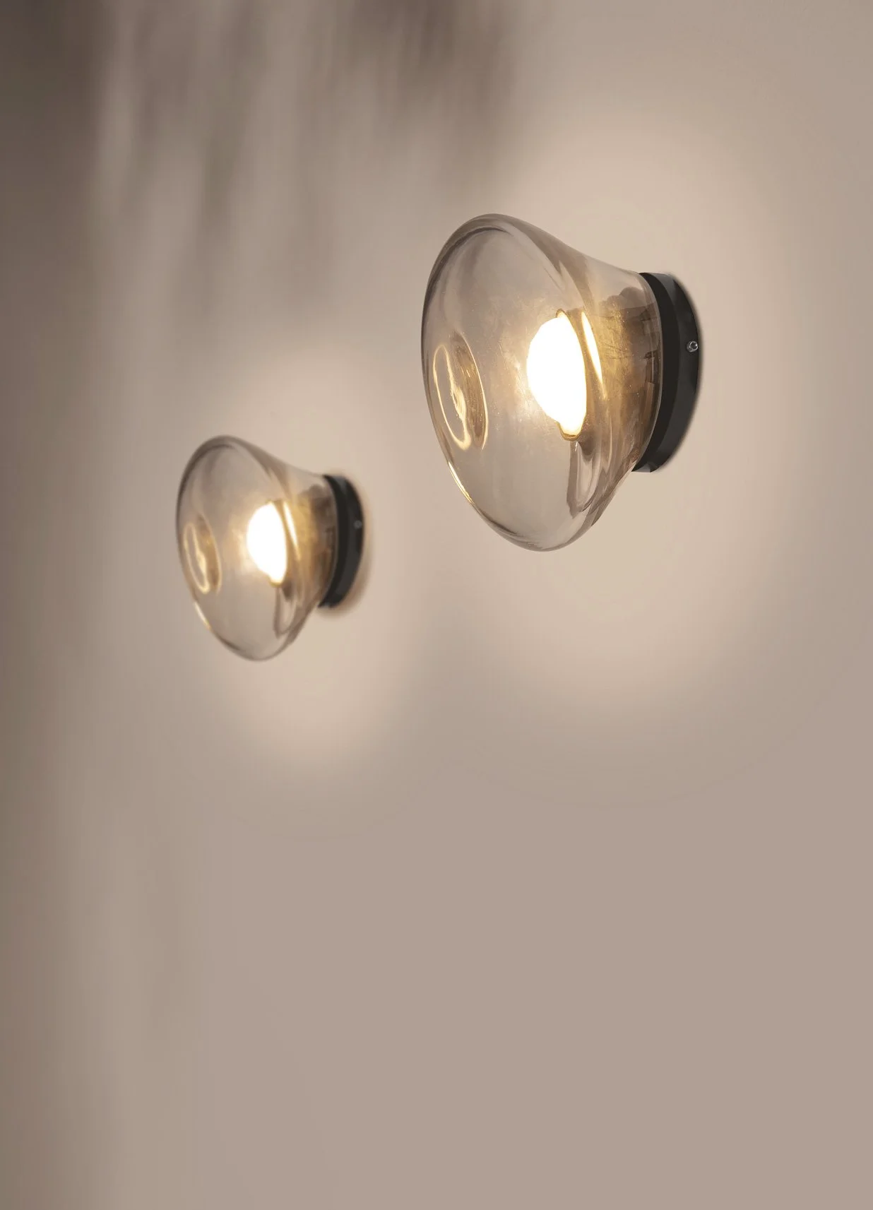 Due applique da parete con vetro trasparente e base nera, accese e agganciate a una parete beige.