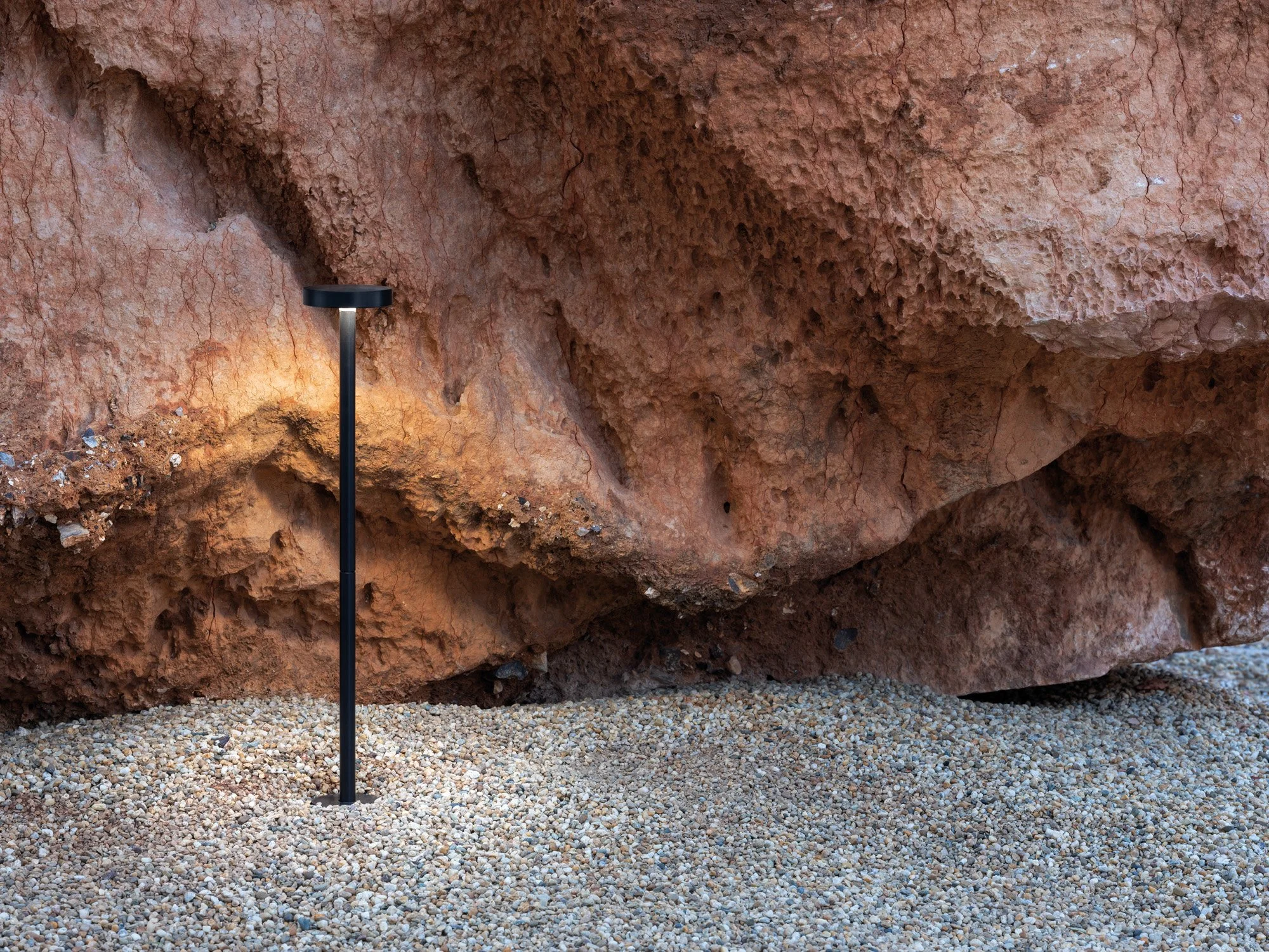 Parete rocciosa rossa con un minimo di terra e sassi sul suolo, illuminata da una luce che viene da sinistra, con un palo di illuminazione nero in primo piano.