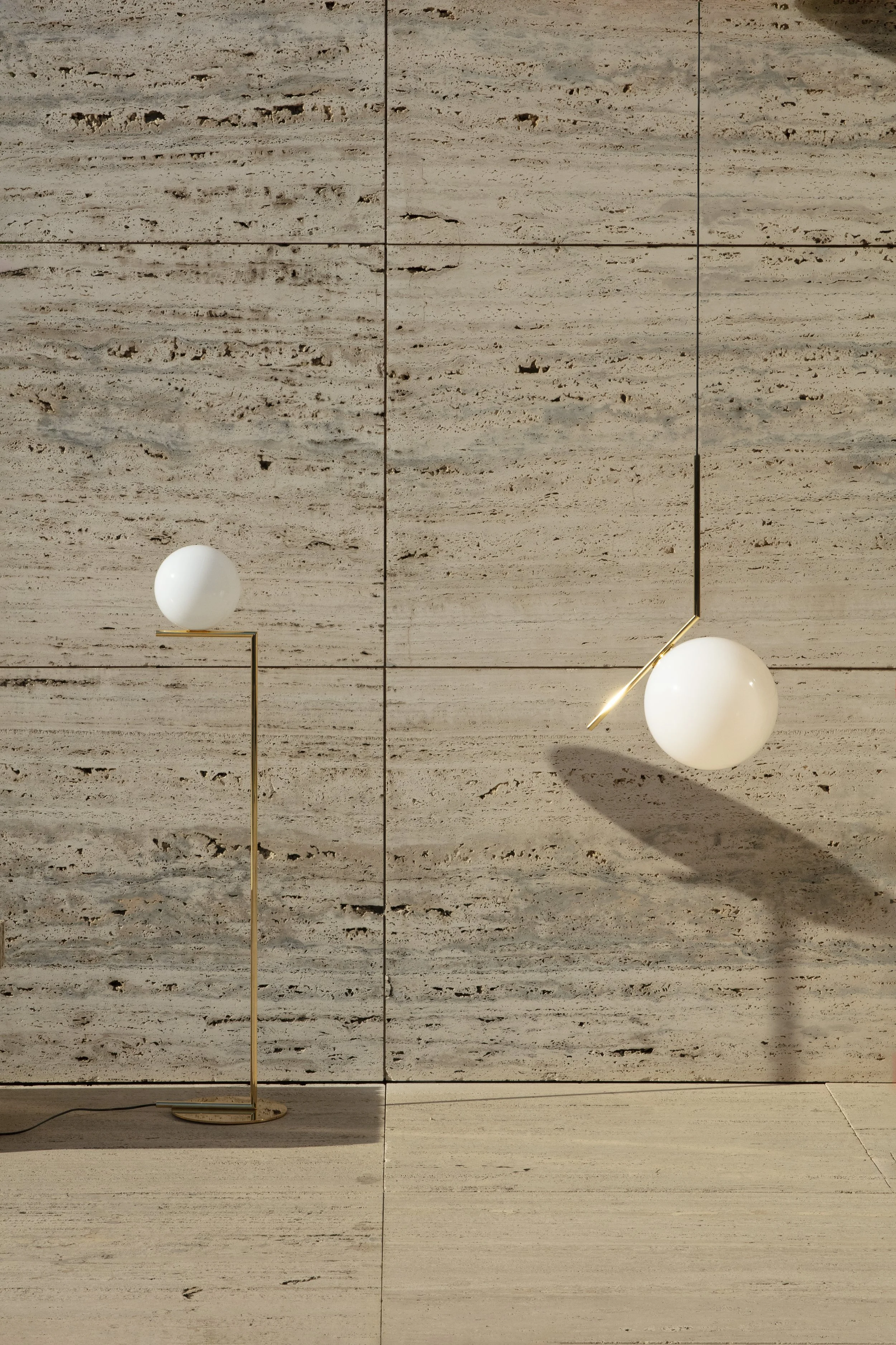 Due lampade da terra con basi rotonde e strutture sottili in metallo dorato, ciascuna con una sfera bianca come paralume, posizionate su una parete di marmo beige con nervature orizzontali.
