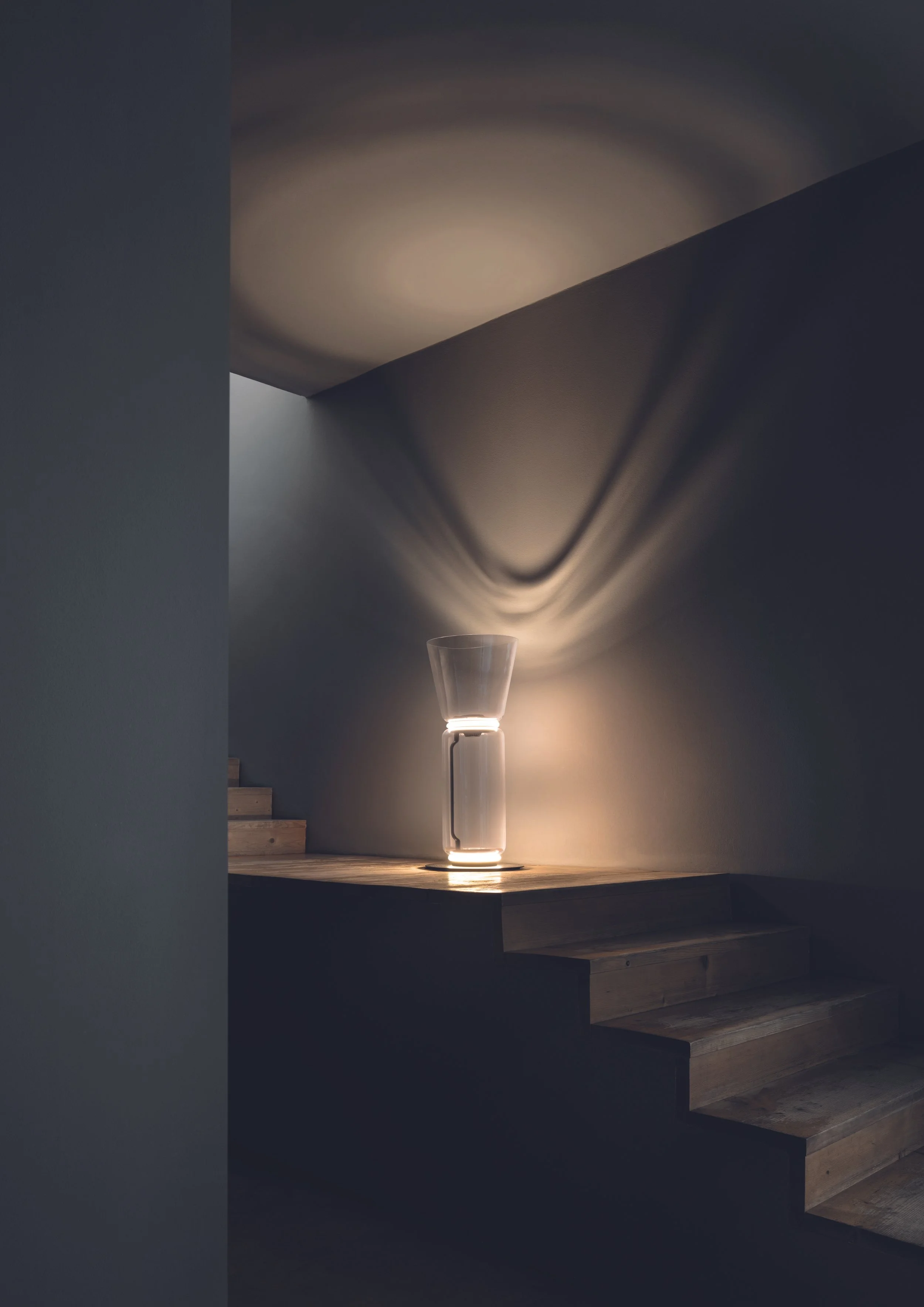 Lampada moderna con finitura trasparente, posizionata su una superficie di legno vicino a una scala di legno, con una luce calda che crea un effetto di ombre sulle pareti circostanti.