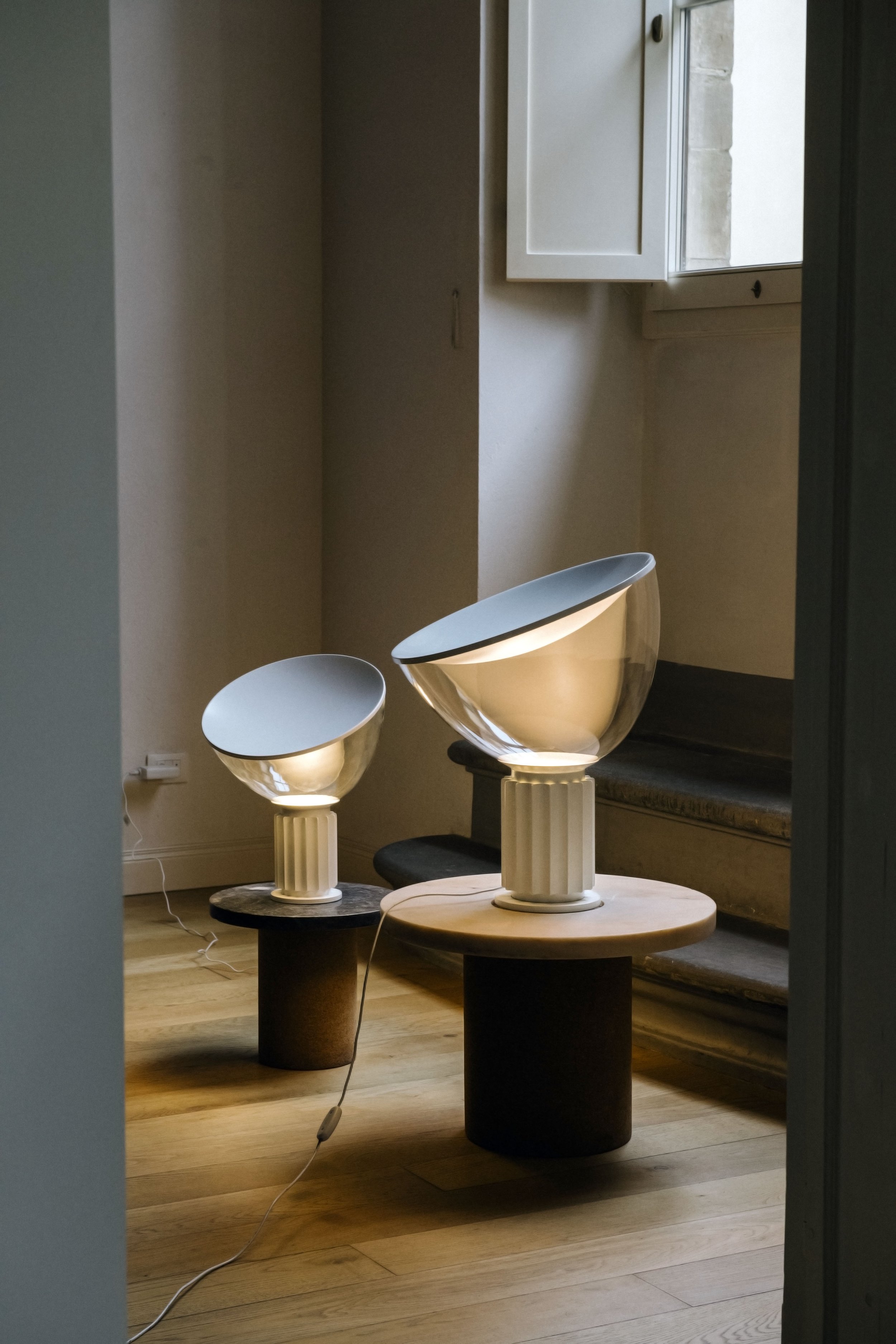 Due lampade da tavolo moderne con base a colonna e paralumi metallici in smalto bianco, posizionate su tavoli di legno, in un ambiente con pareti chiare e pavimento in legno, vicino a una finestra con persiana, illuminazione soffusa.