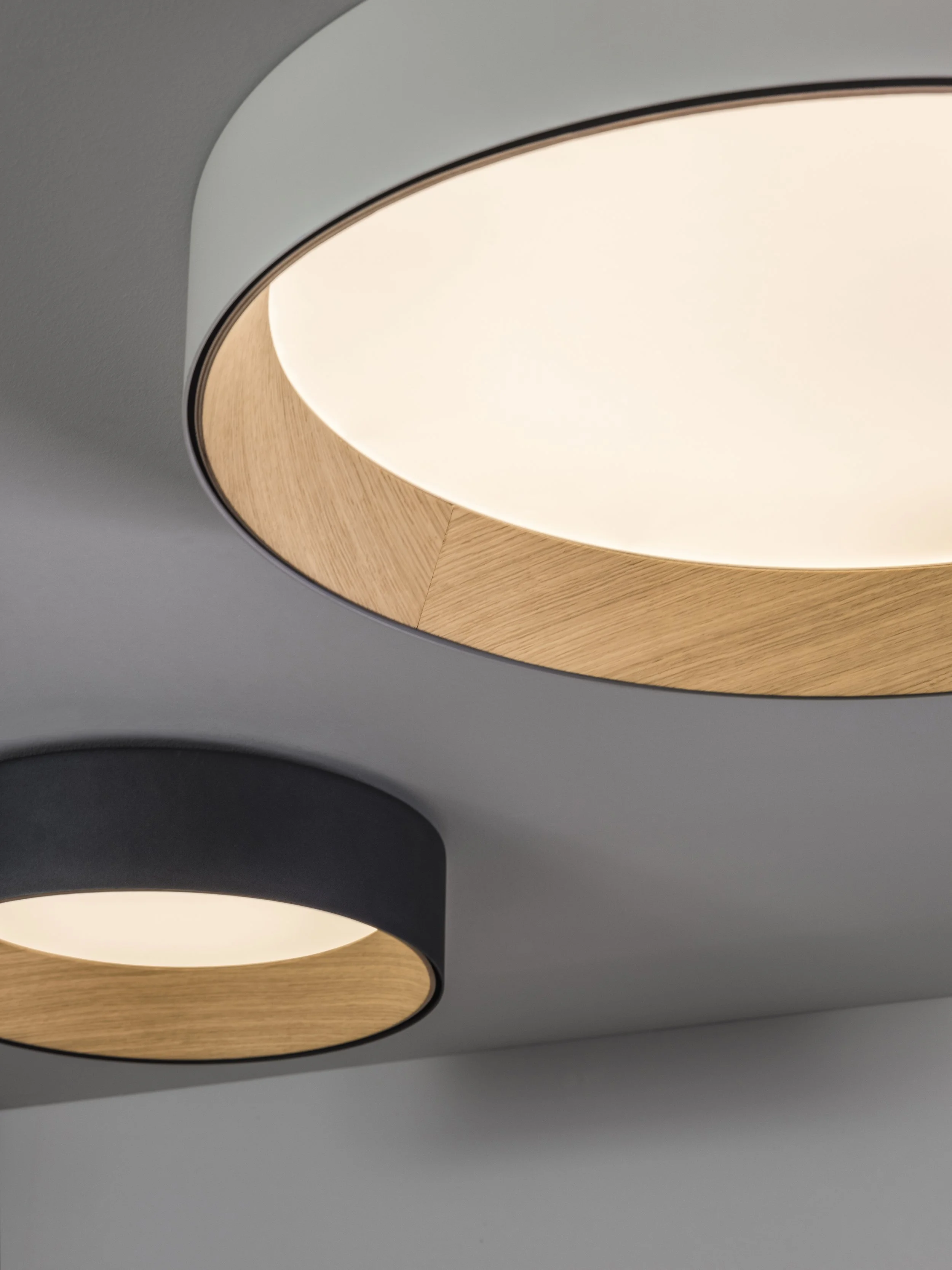 Particolare di lampade moderne con diffusori circolari in materiale bianco e supporti in legno, installate su un soffitto grigio.