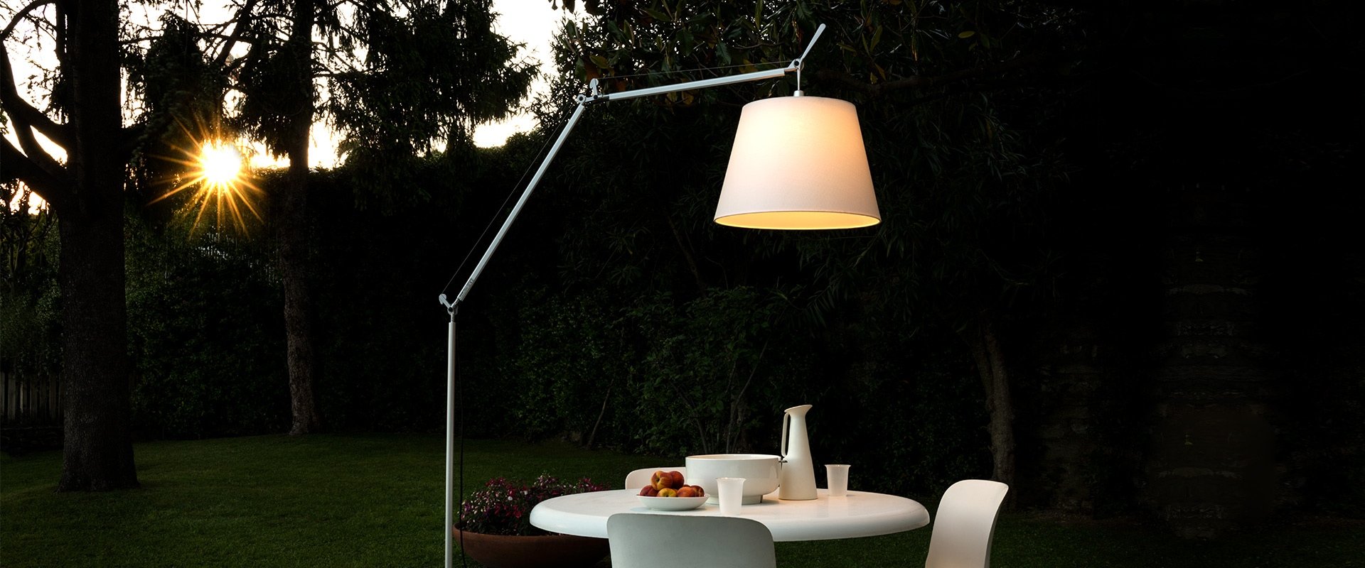 Tavolo da esterno con ciotola di frutta e stoviglie, illuminato da una lampada sospesa, in un giardino al tramonto.