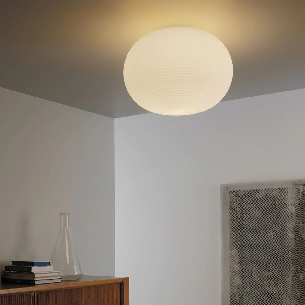 Lampada da soffitto rotonda bianca con luce calda, pareti bianche, scaffale con libri e un vaso di vetro, elemento decorativo a parete con motivo geometrico.