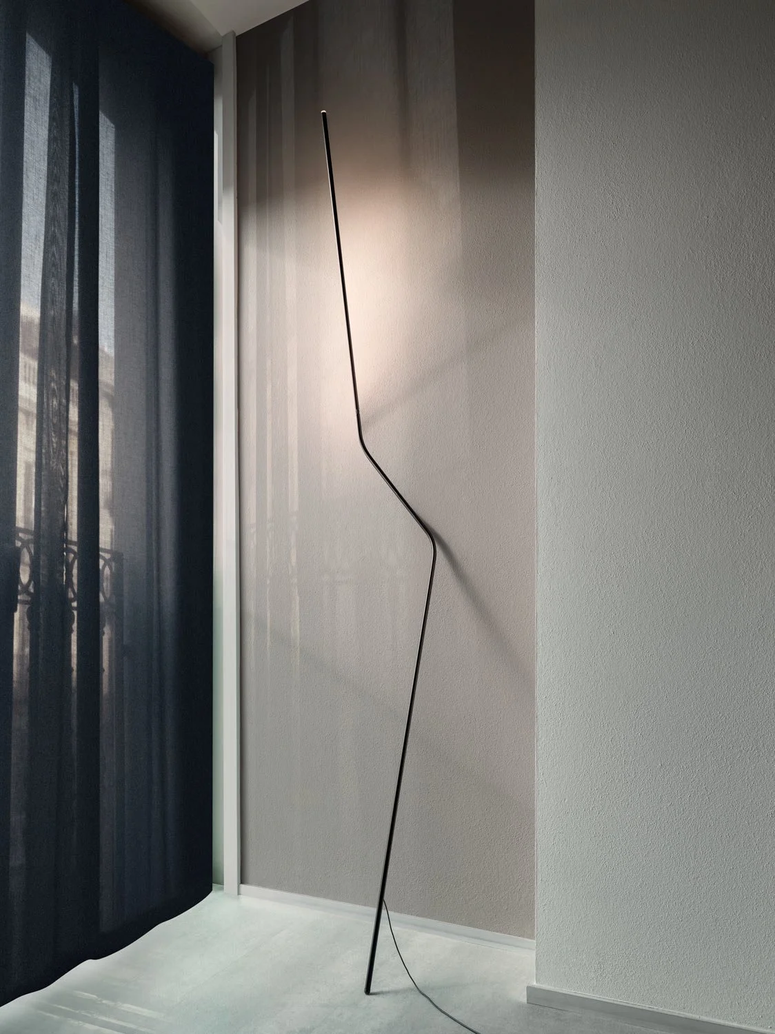Lampada da parete moderna e minimalista con design sottile e lineare, installata su una parete di colore chiaro vicino a una finestra con tende nere.