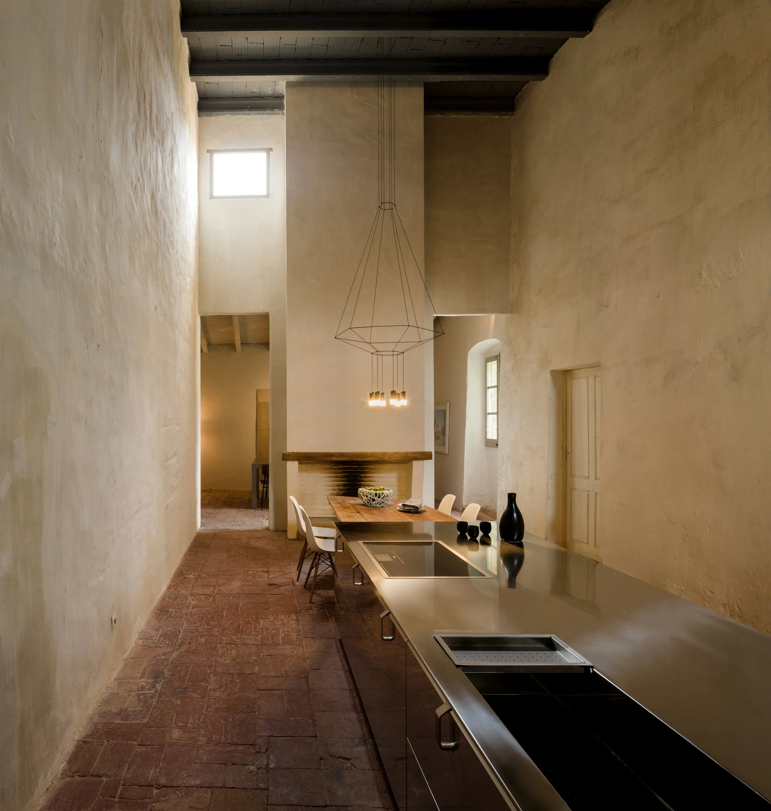 Interno di una stanza con pareti di colore beige, soffitto con travi di legno e un lampadario sospeso geometricamente, cucina moderna con piano cottura a induzione, tavolo e sedie, grandi finestre e porte in legno in uno stile rustico.