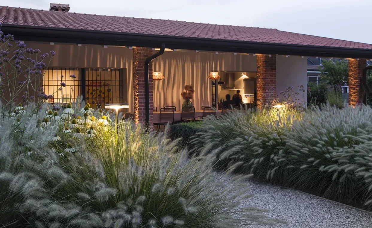 Casa con veranda illuminata di sera, circondata da piante ornamentali e fiori