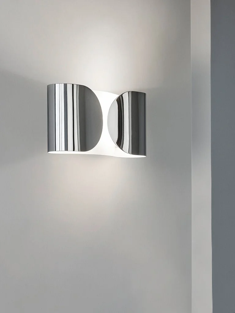 Specchio da parete con design moderno e finitura metallica, illuminato da luce soffusa su una parete bianca.