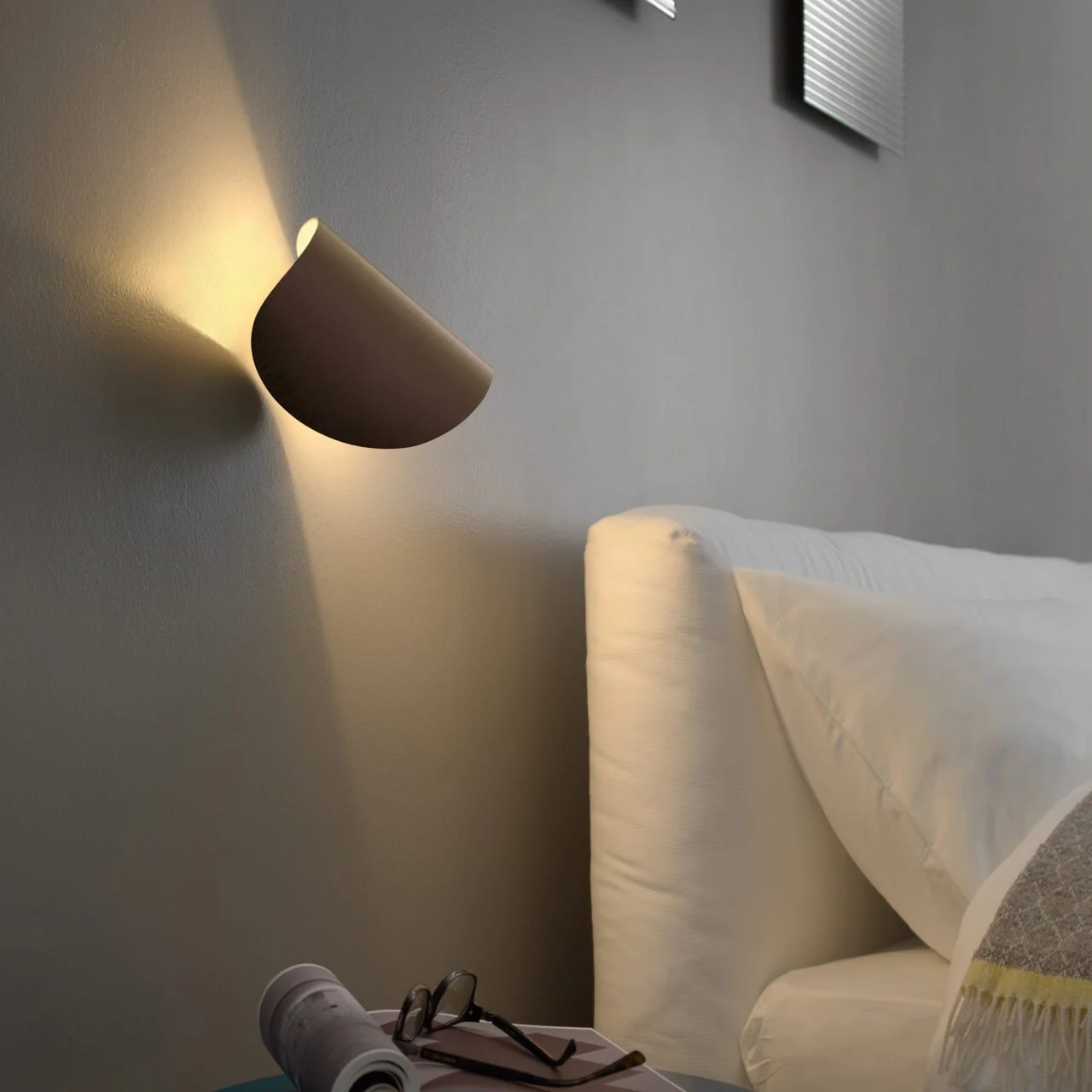 L'immagine mostra una stanza con una parete grigia, con una lampada da muro accesa, una testa di letto imbottita e un comodino con occhiali, un rotolo di carta e altri oggetti