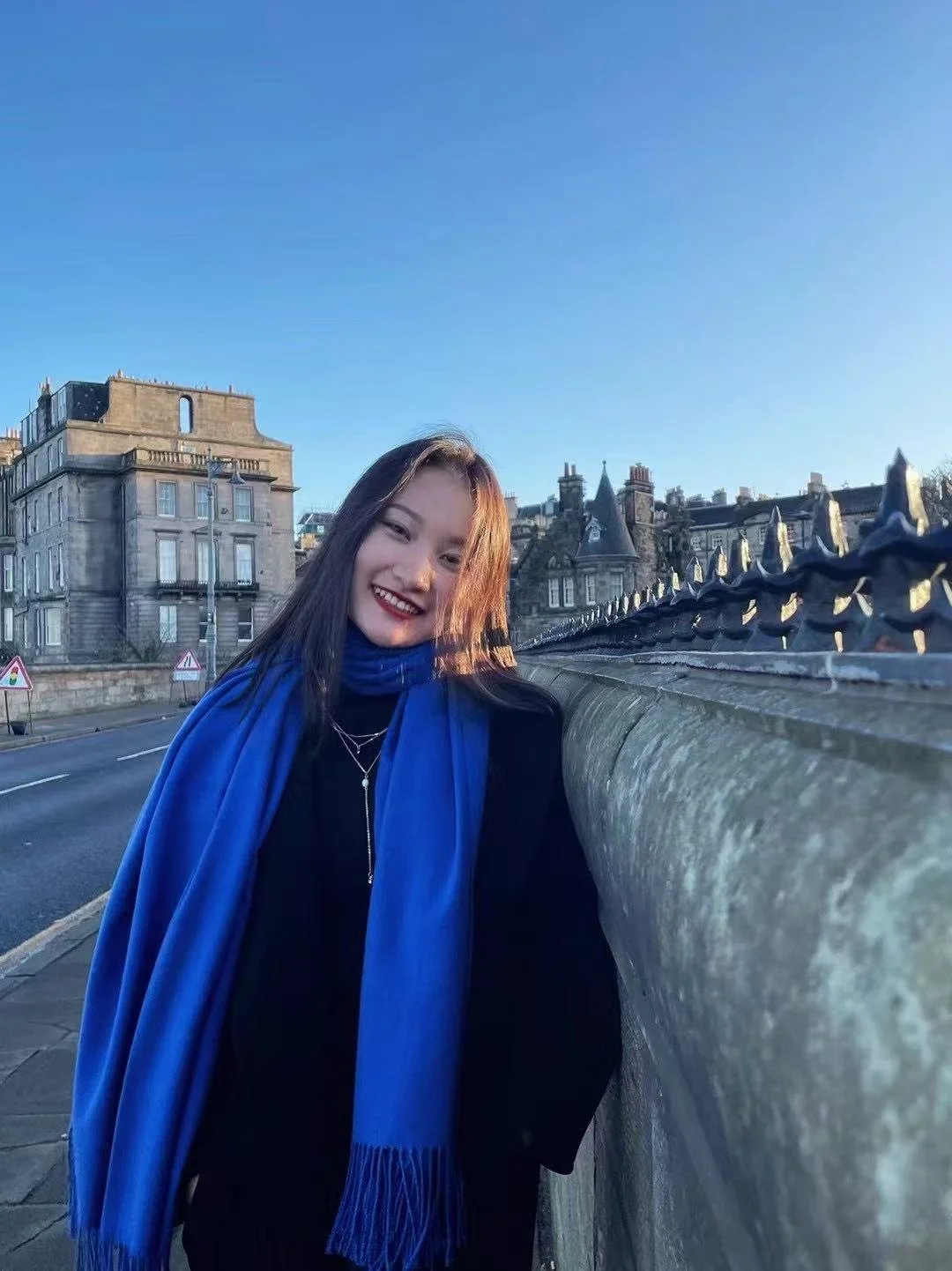 A young woman wearing a black coat and a bright blue scarf, smiling and standing next to a stone bridge railing on a clear sunny day in a city with historic buildings.