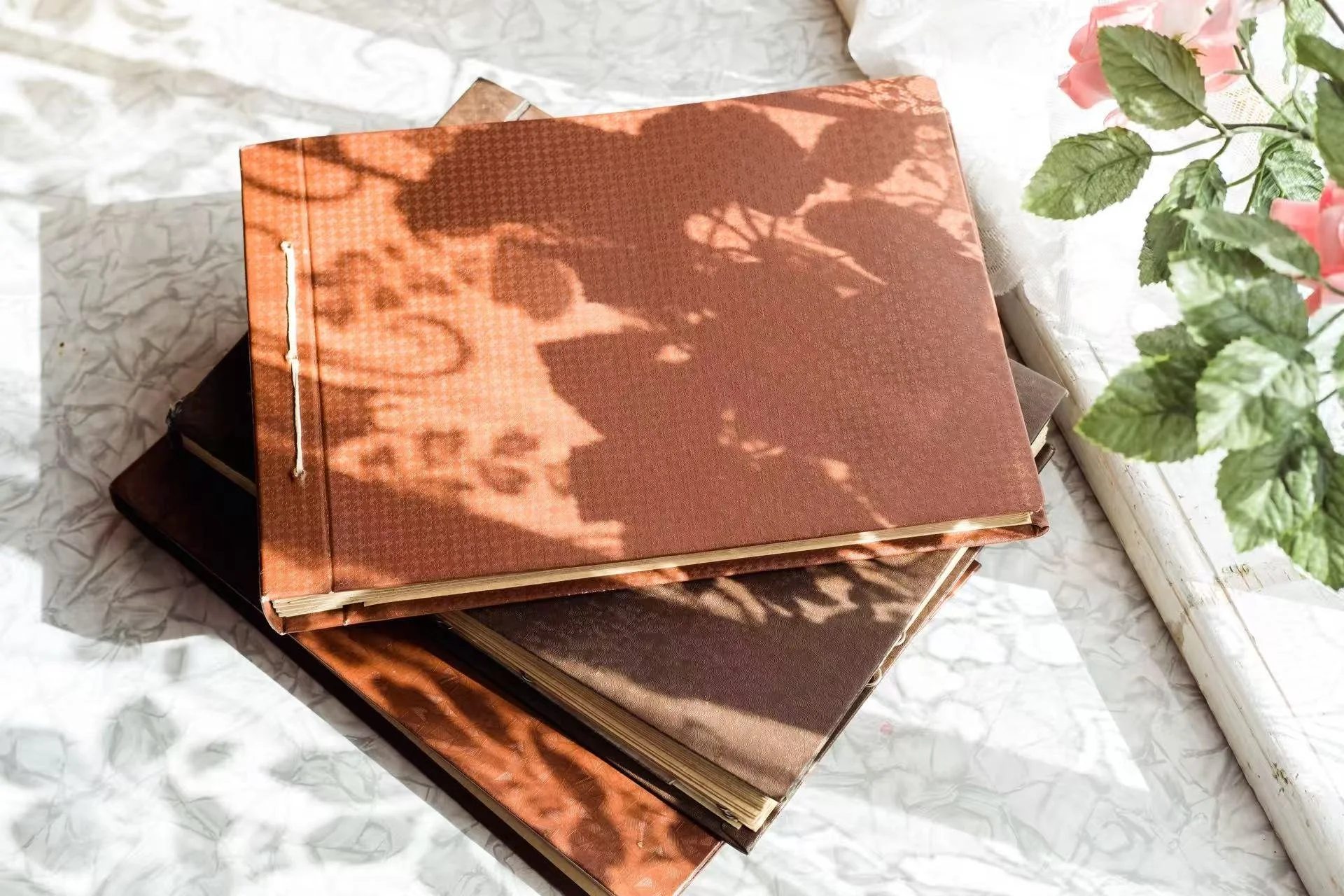 A pile of closed books with textured covers, with sunlight casting shadows of leaves on the top book, sitting on white crumpled paper next to a white windowsill with green leaves and pink flowers.
