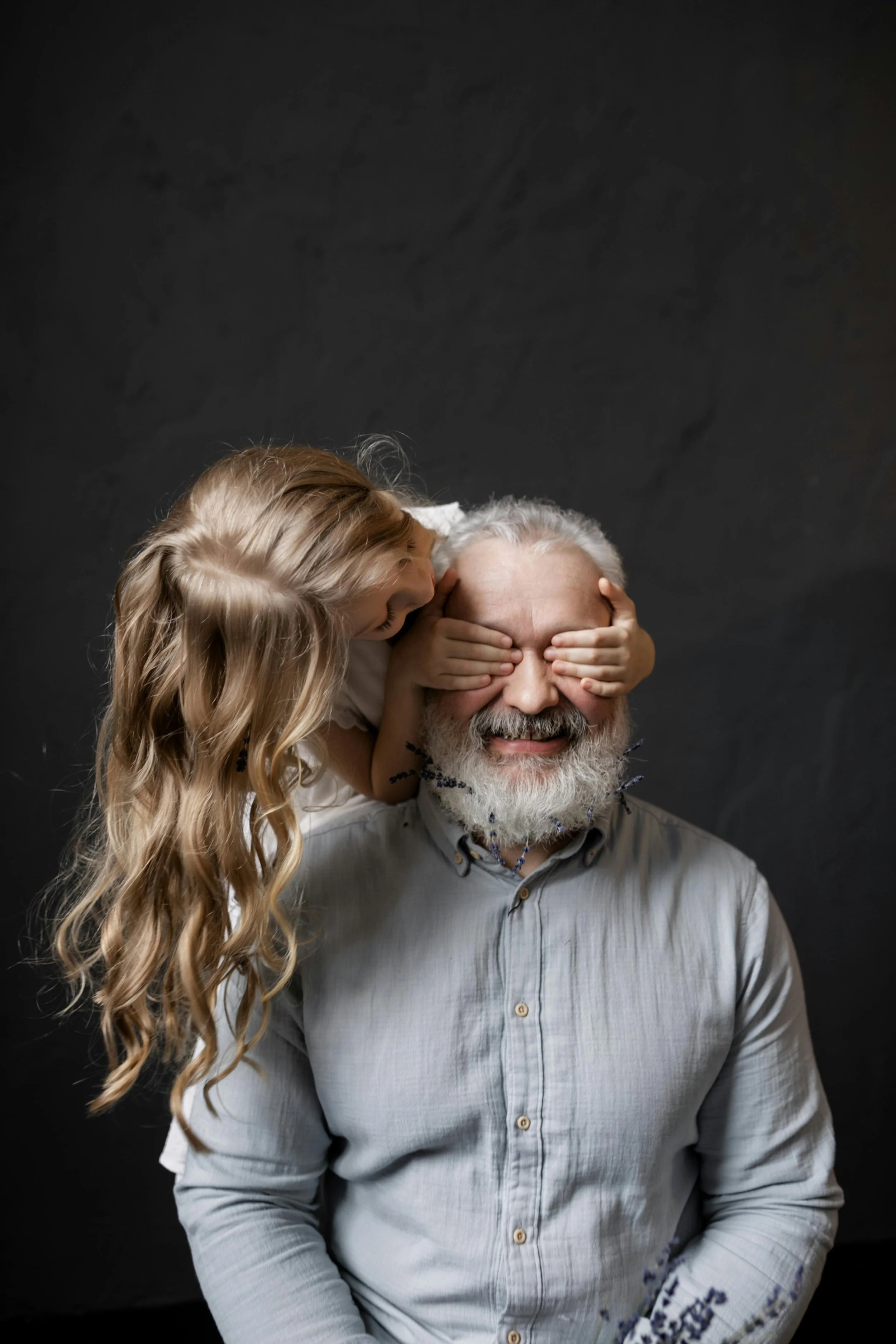 A young girl with long, wavy blonde hair whispering into an older man's ear while her hands cover his eyes, with the man smiling and wearing a gray button-up shirt, against a dark backdrop.