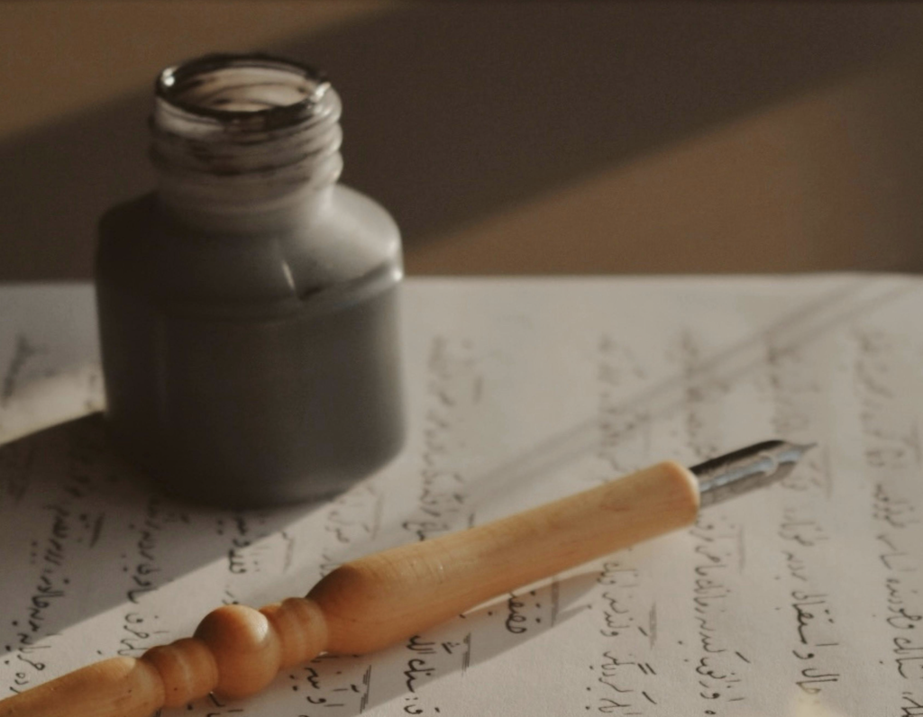 An open ink bottle and a calligraphy pen with a wooden handle resting on handwritten paper.
