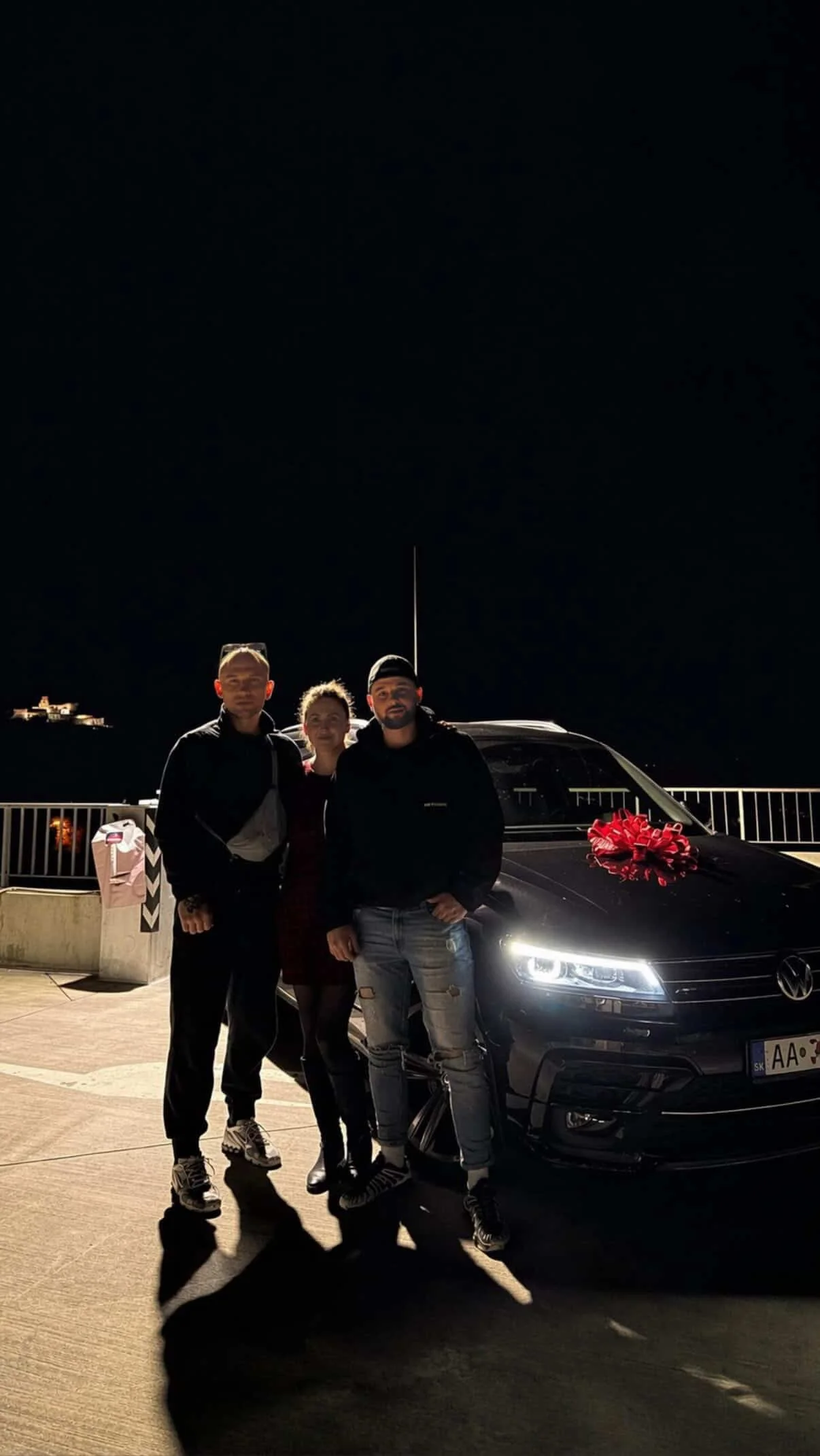 Three people standing together at night next to a black car with a large red bow on the hood, on a rooftop or parking area with a railing and city or landscape in the background.