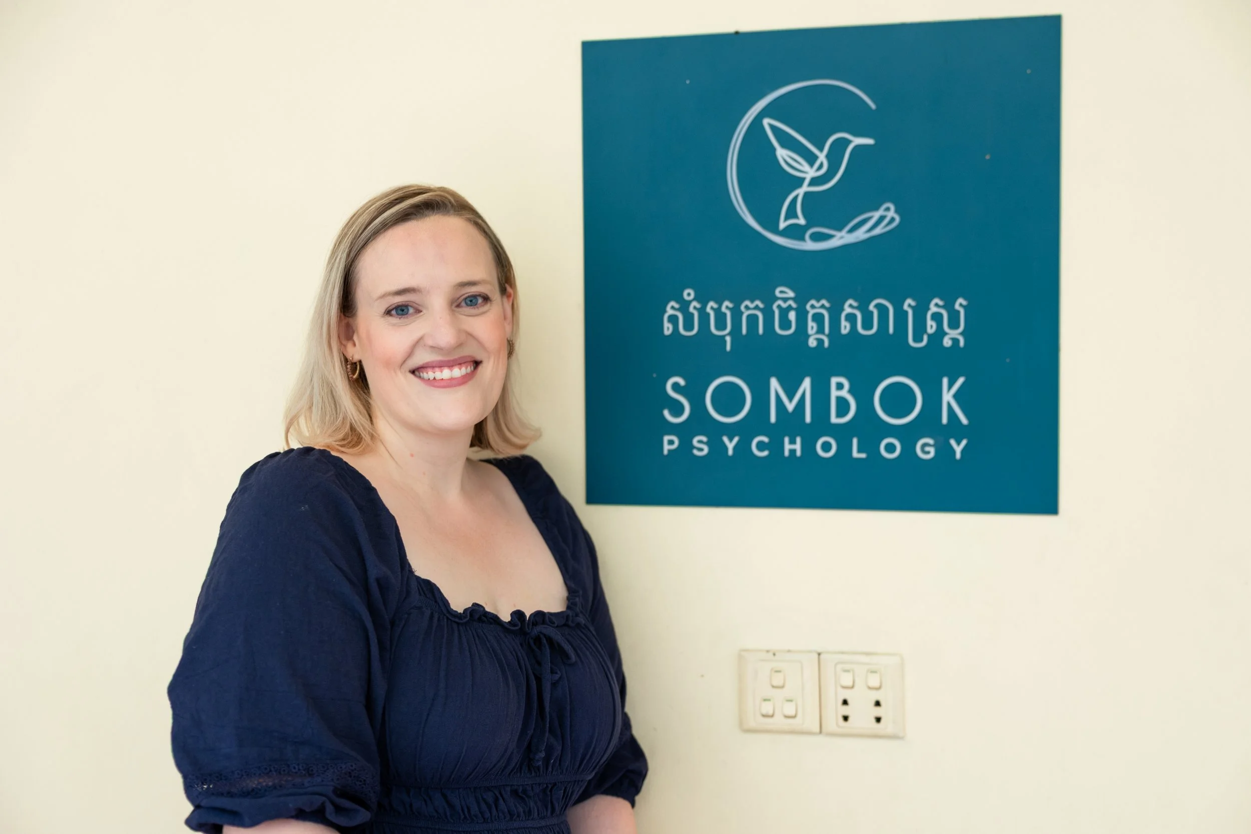 Smiling woman standing next to a blue sign that reads 'Sombok Psychology' with a hummingbird logo, on a cream wall background.
