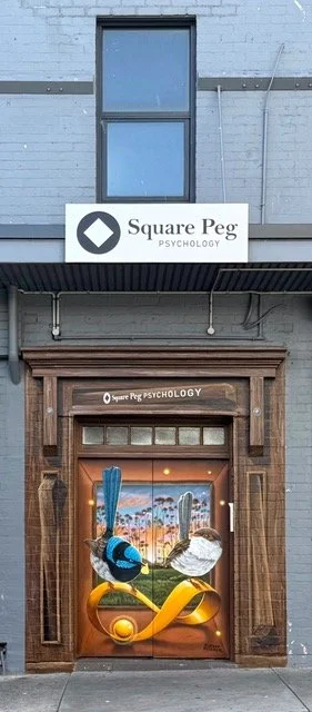 Storefront with a sign reading 'Square Peg Psychology' above a decorated door featuring a blue bird, a white bird, and a yellow infinity symbol.