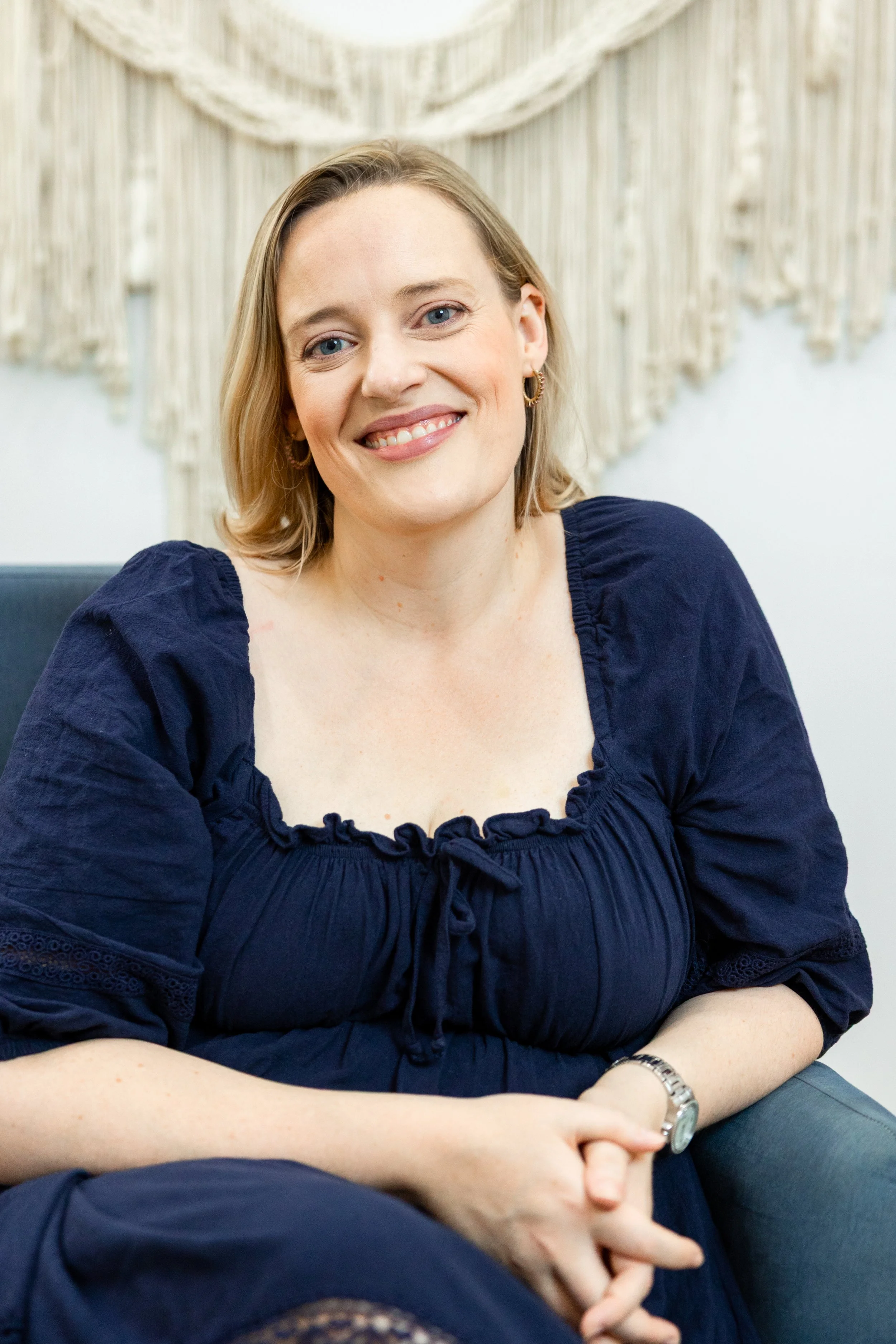 A smiling woman with blonde hair wearing a navy blue top with ruffled sleeves and gold hoop earrings, sitting with her arms crossed against a light-colored macramé wall hanging.
