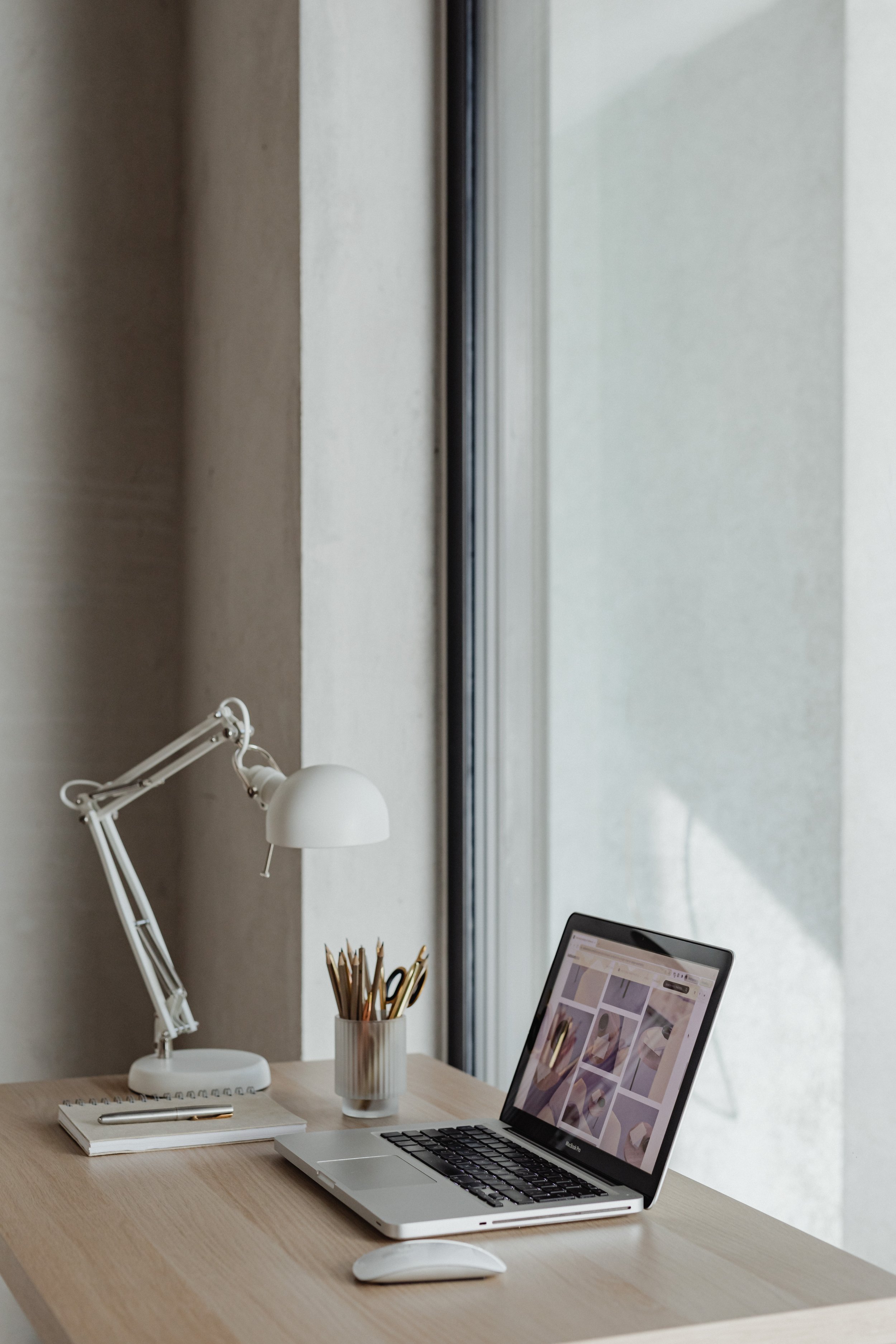 A tidy workspace with a wooden desk, a white adjustable desk lamp, a potted plant with pencils, a notebook, a pen, a laptop displaying a collage of images, and a wireless mouse, set in front of a large window with a beige wall behind.
