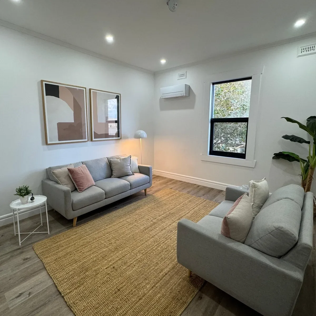 Living room with two light gray sofas, a wooden floor, a beige woven rug, a small white side table with a potted plant, framed abstract art on the wall, a floor lamp, and a window with a view of trees.