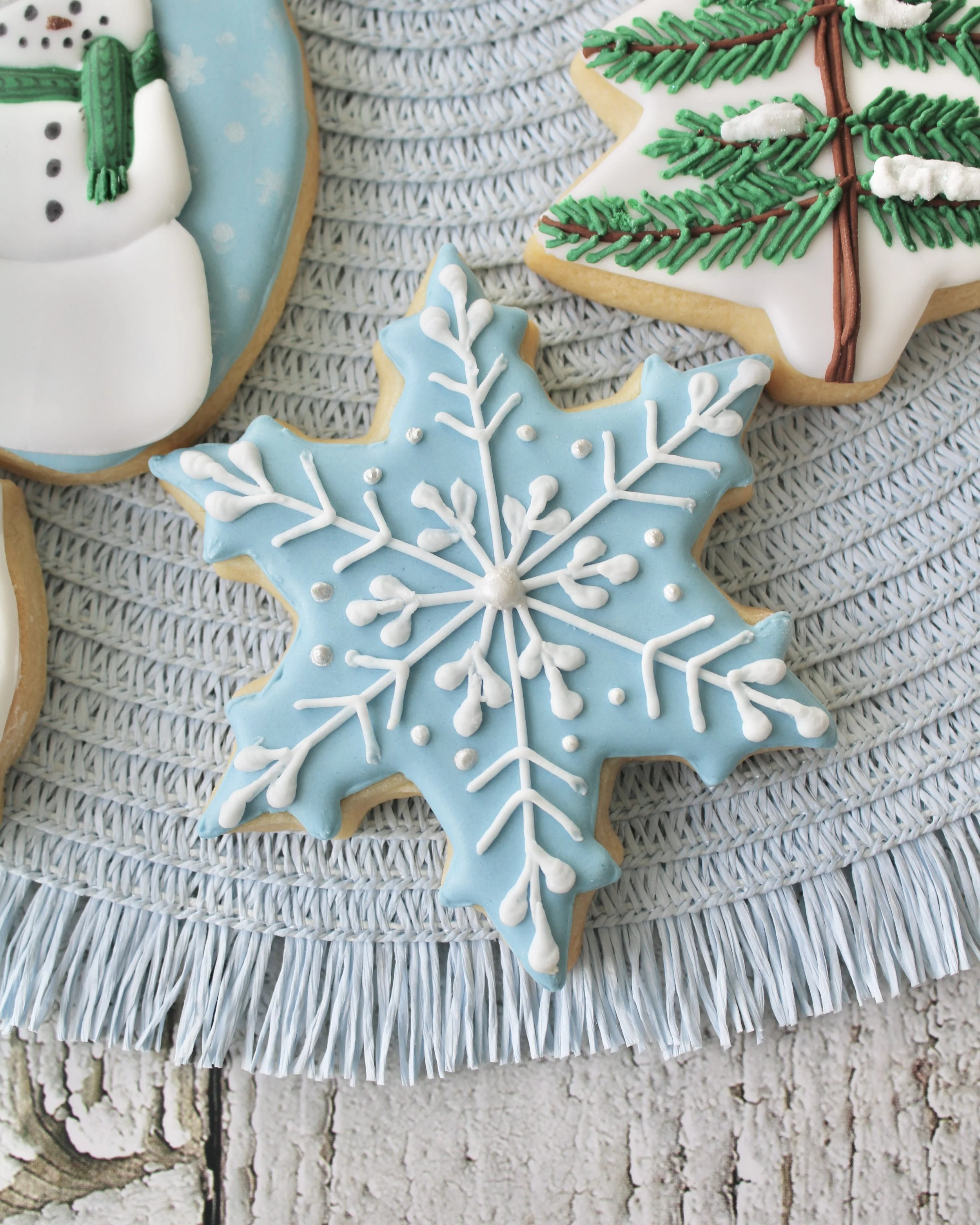 Close-up pale blue snowflake cookie with white icing details, styled on pale blue placemat.