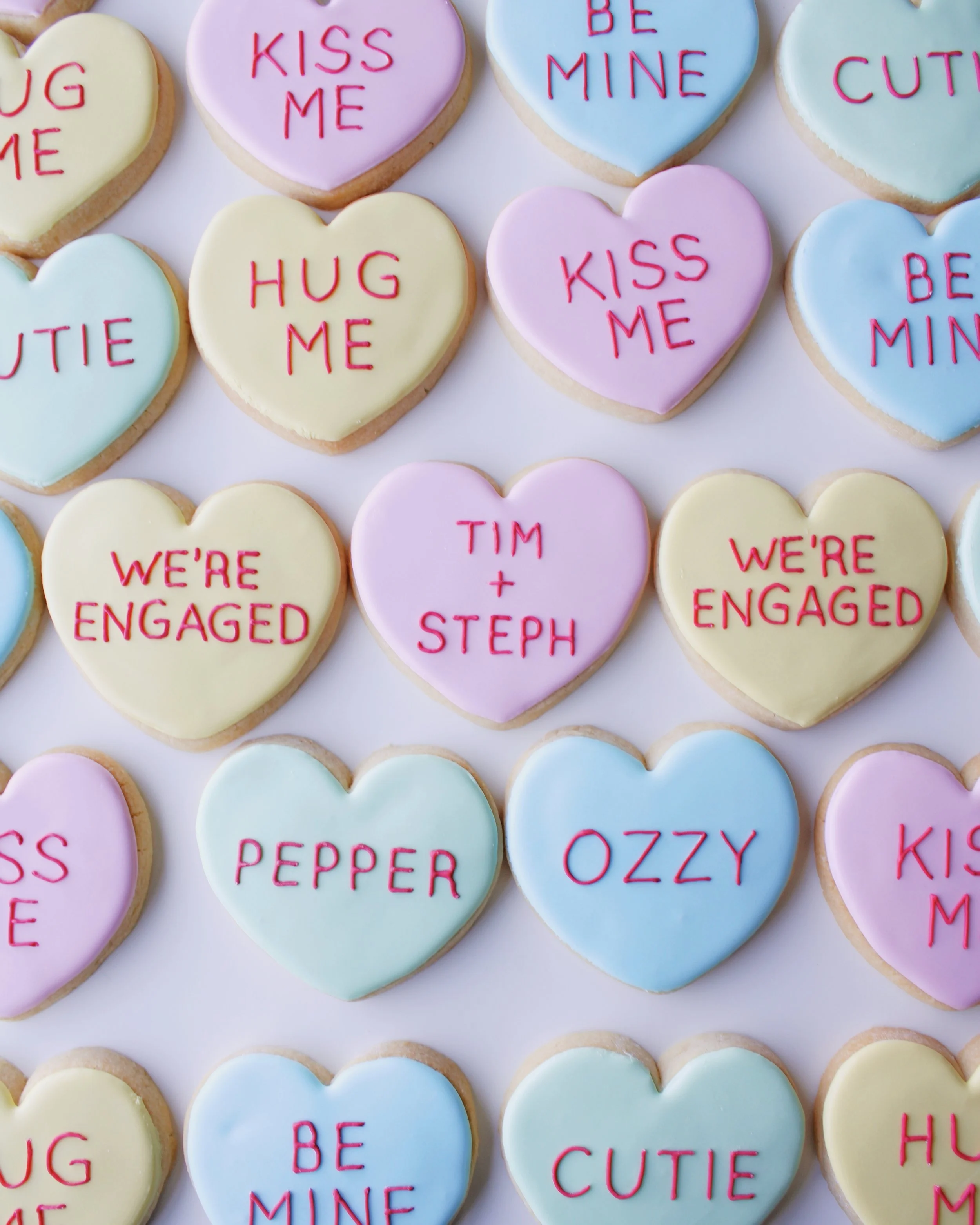 Heart-shaped cookies decorated like conversation heart candies in pastel coloured icing, with red text messages such as “hug me", “kiss me,” “be mine,” “Tim + Steph,” and “we’re engaged.”