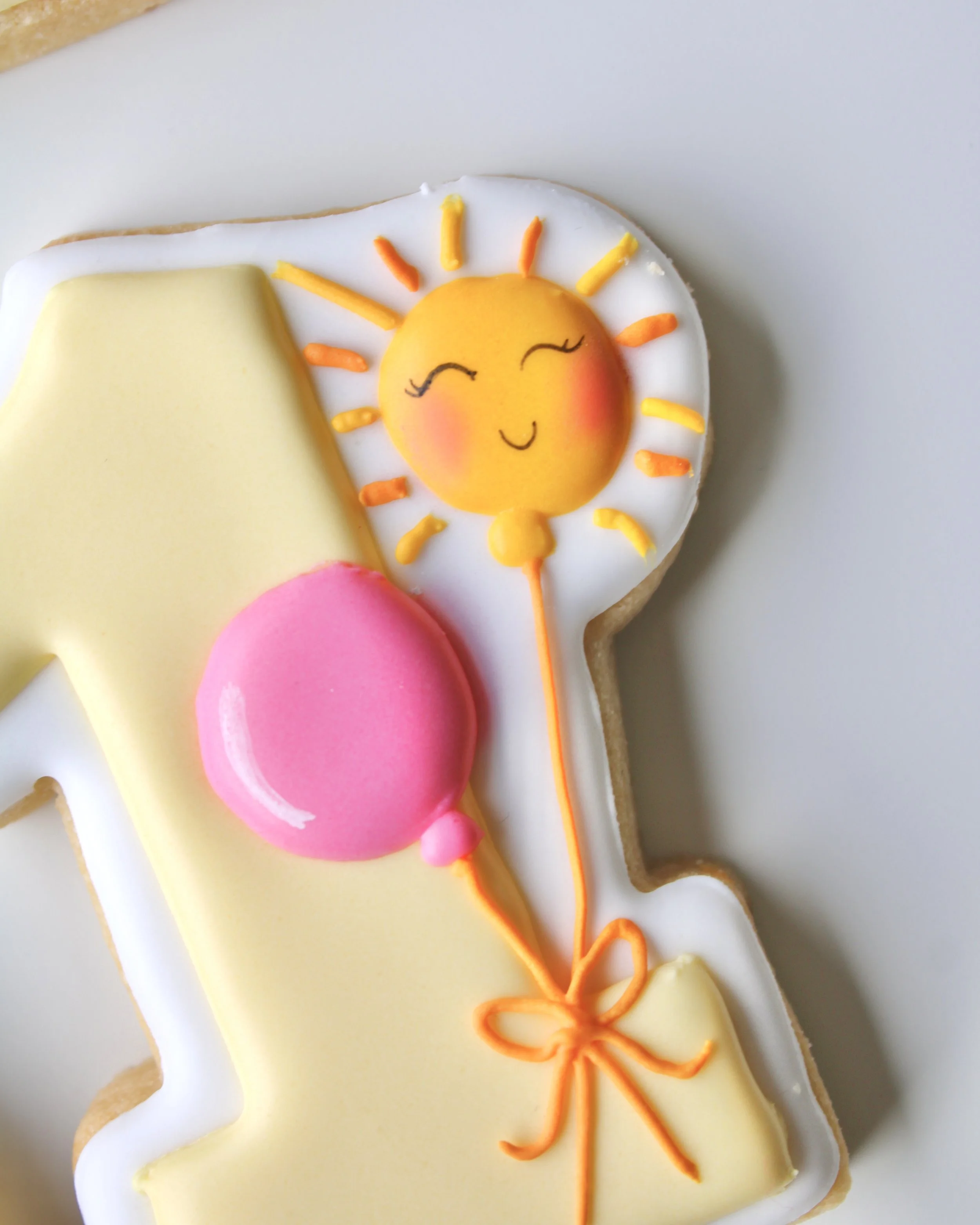 Close-up of a number 1 cookie iced with butter yellow icing, decorated with a pink balloon and a sun-shaped balloon design.