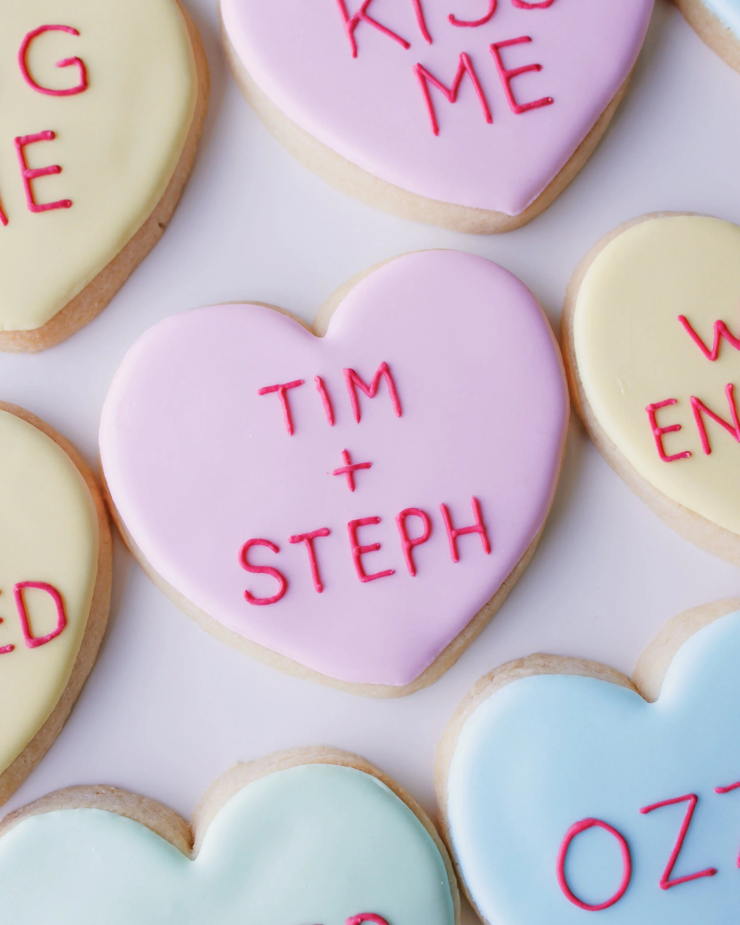 Close-up of a heart-shaped cookie decorated like a conversation heart candy in pastel pink icing with red personalised text reading “Tim + Steph.”