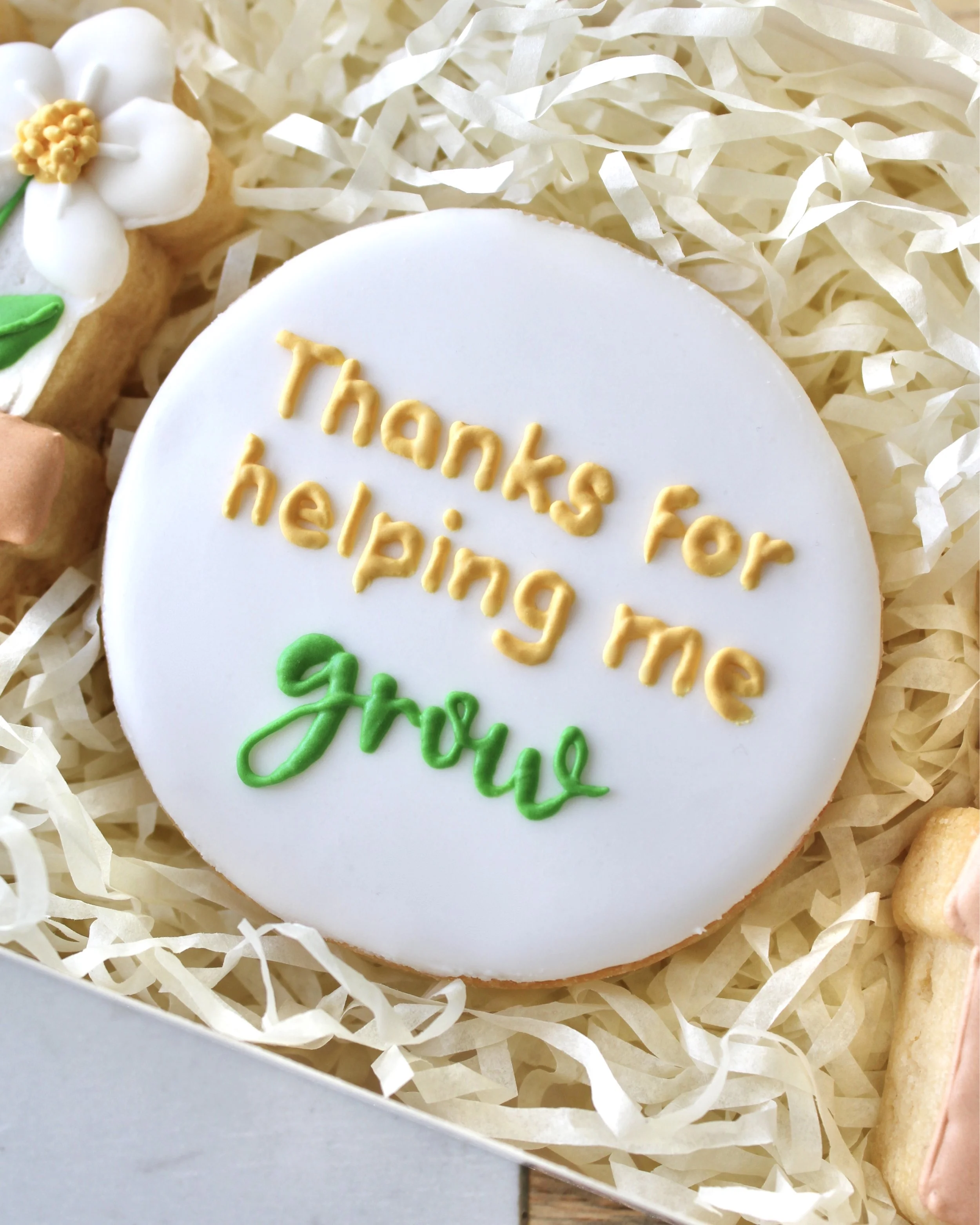 Close-up of a round white cookie piped with “thank you for helping me grow” in yellow and green royal icing.