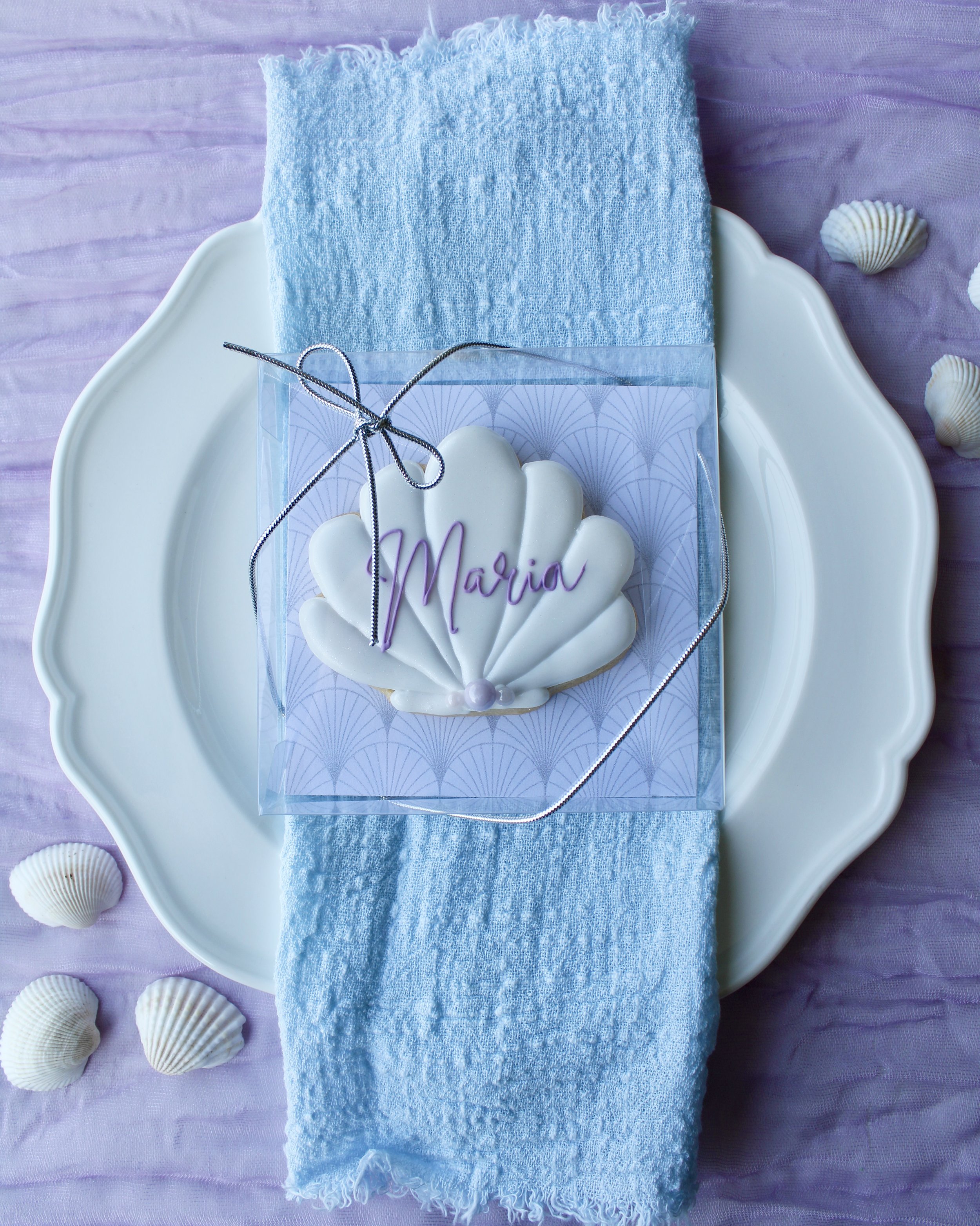White seashell cookie with “Maria” in purple cursive icing, in a clear box tied with silver cord bow, on a white vintage plate over blue linen and purple crinkled fabric with seashells.