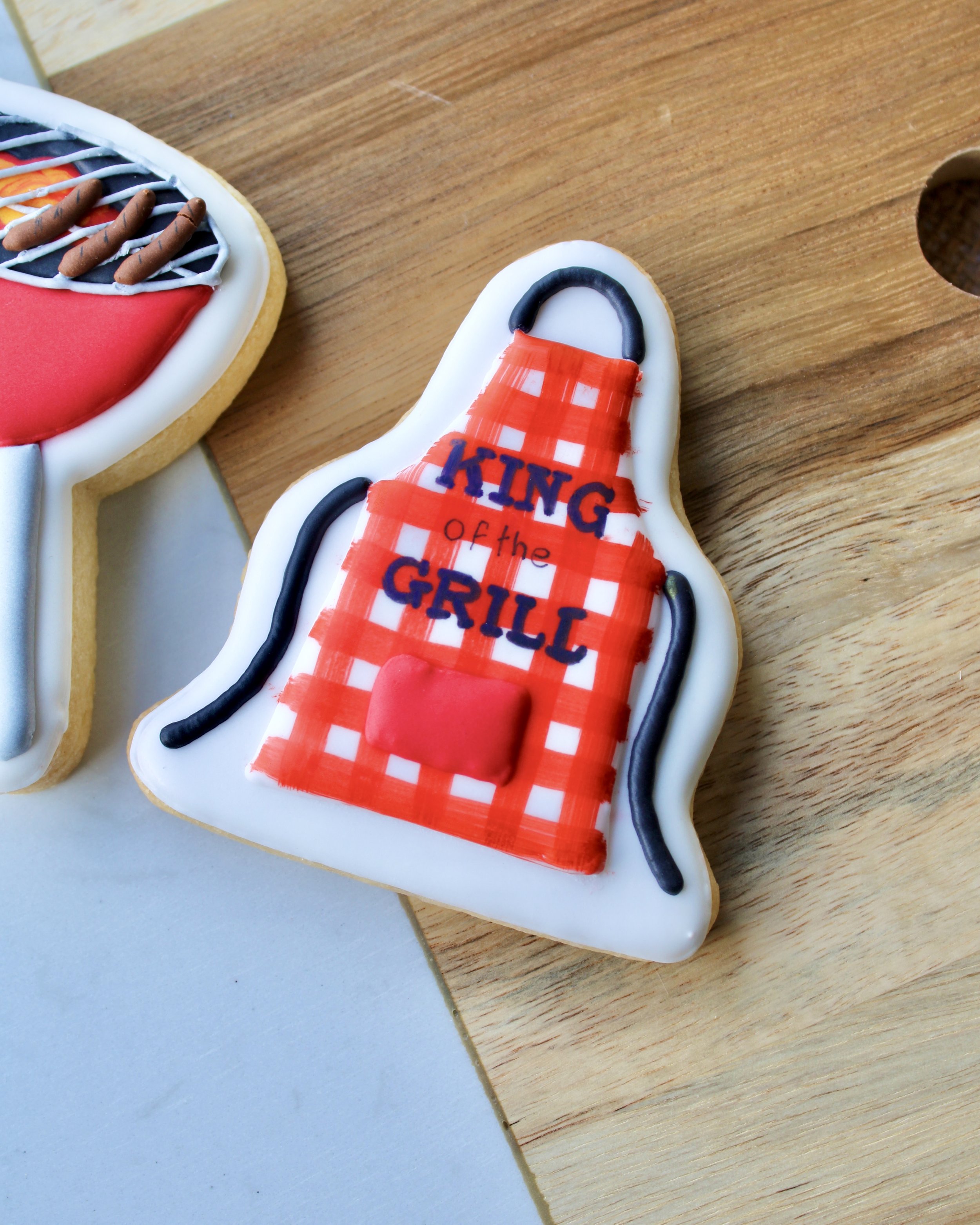 Close up of Father's Day cookie featuring a red and white gingham patterned apron with "King of the Grill" written on it, styled on a wooden board.