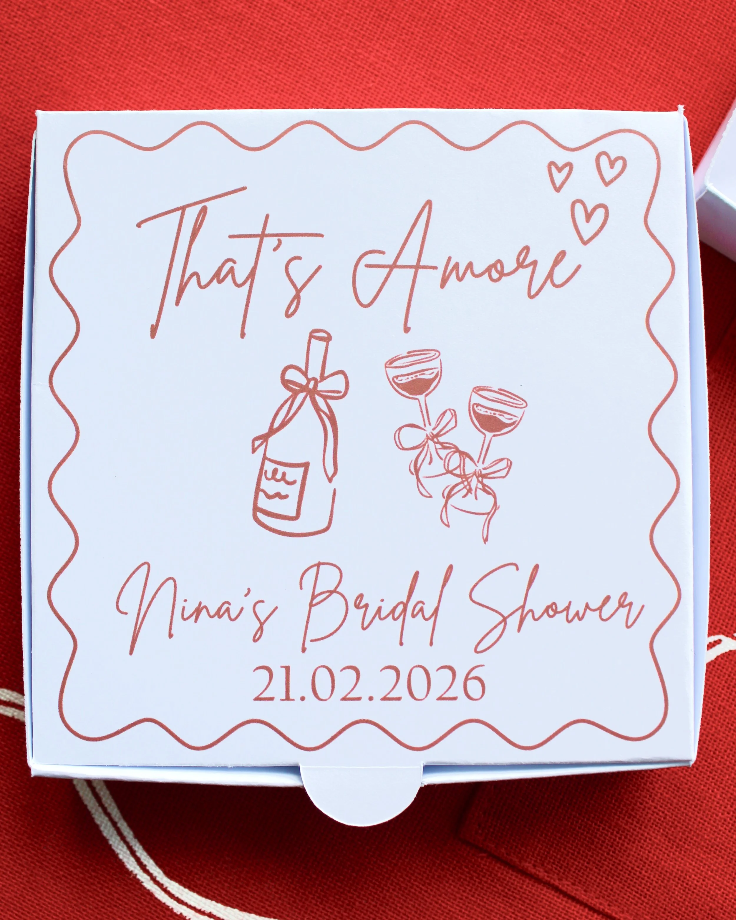 Close up of mini pizza cookie box reading “That’s Amore” with the couple’s names and wedding date, styled on red fabric.
