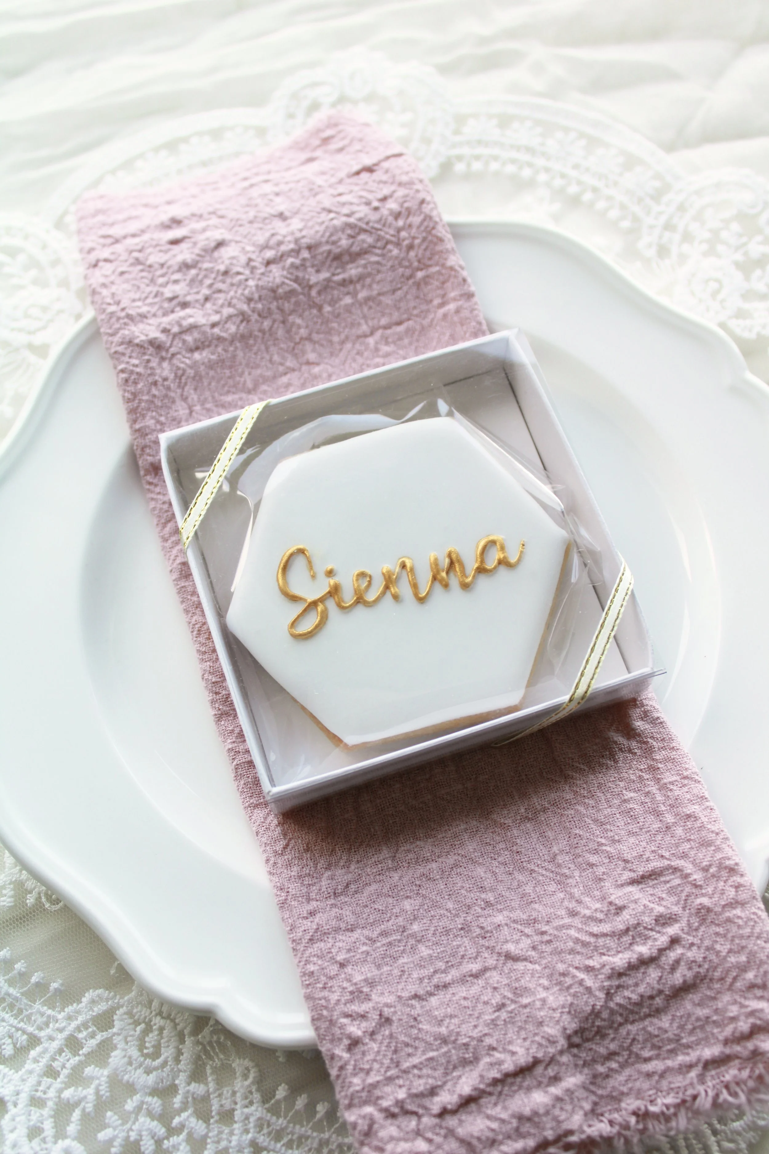 Hexagon place card sugar cookie iced in white royal icing with personalised name “Sienna” in gold script, handmade royal icing cookie styled on a blush napkin and vintage plate with lace detail.