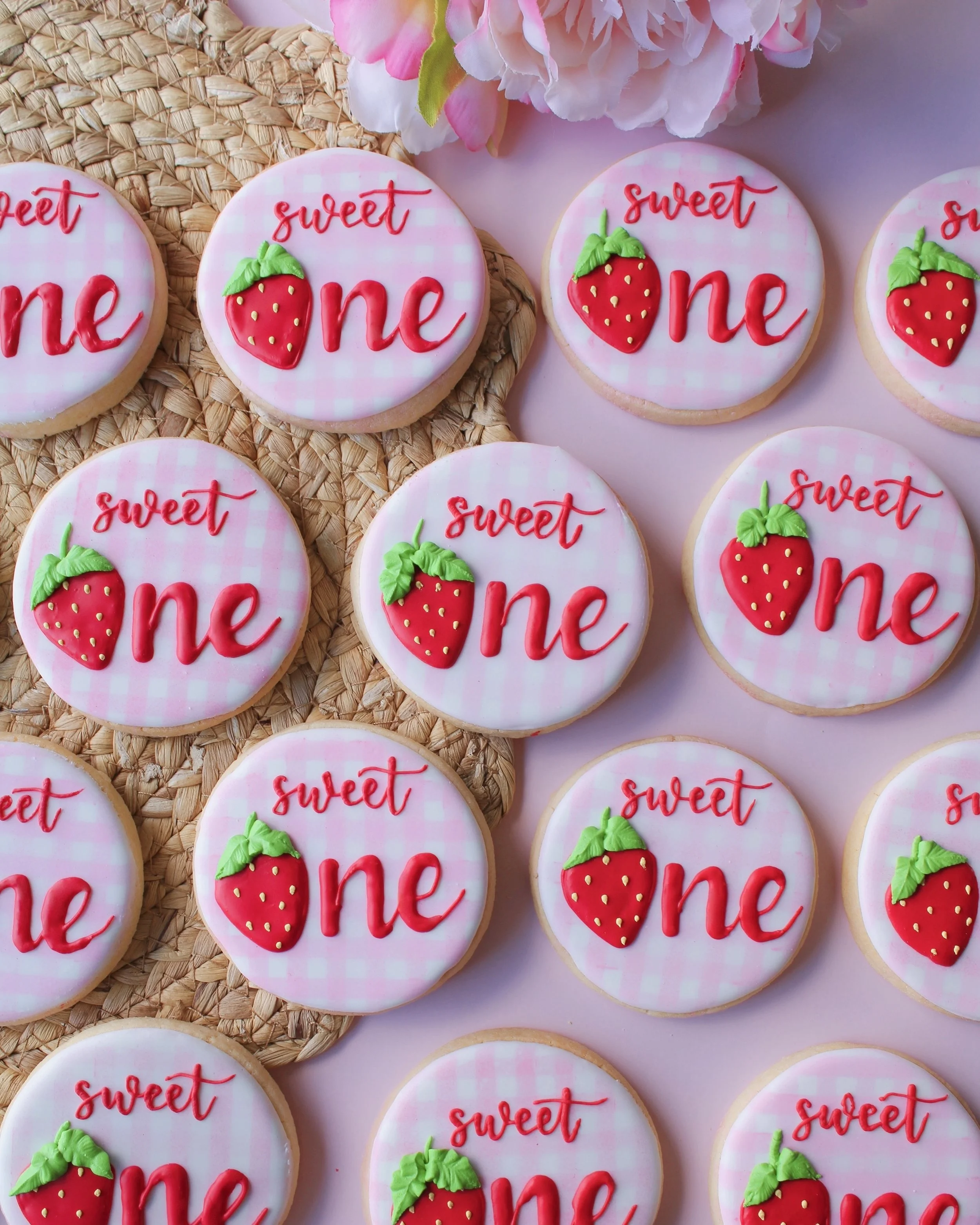 Sweet One birthday sugar cookies with pink and white gingham design, featuring the word “one” in red cursive with a strawberry replacing the “o”.