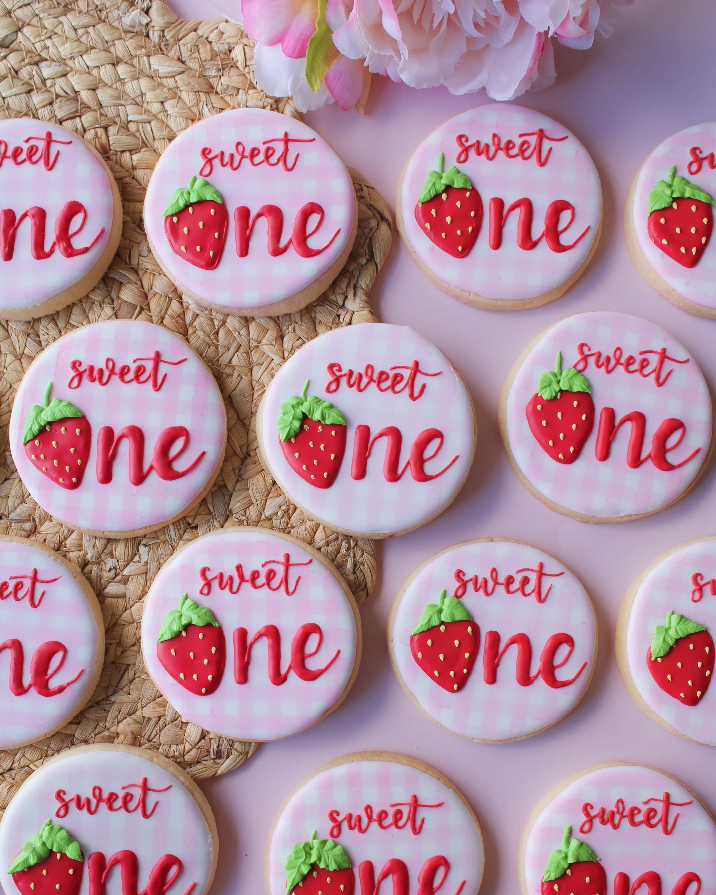 Sweet One birthday sugar cookies with pink and white gingham design, featuring the word “one” in red cursive with a strawberry replacing the “o”.