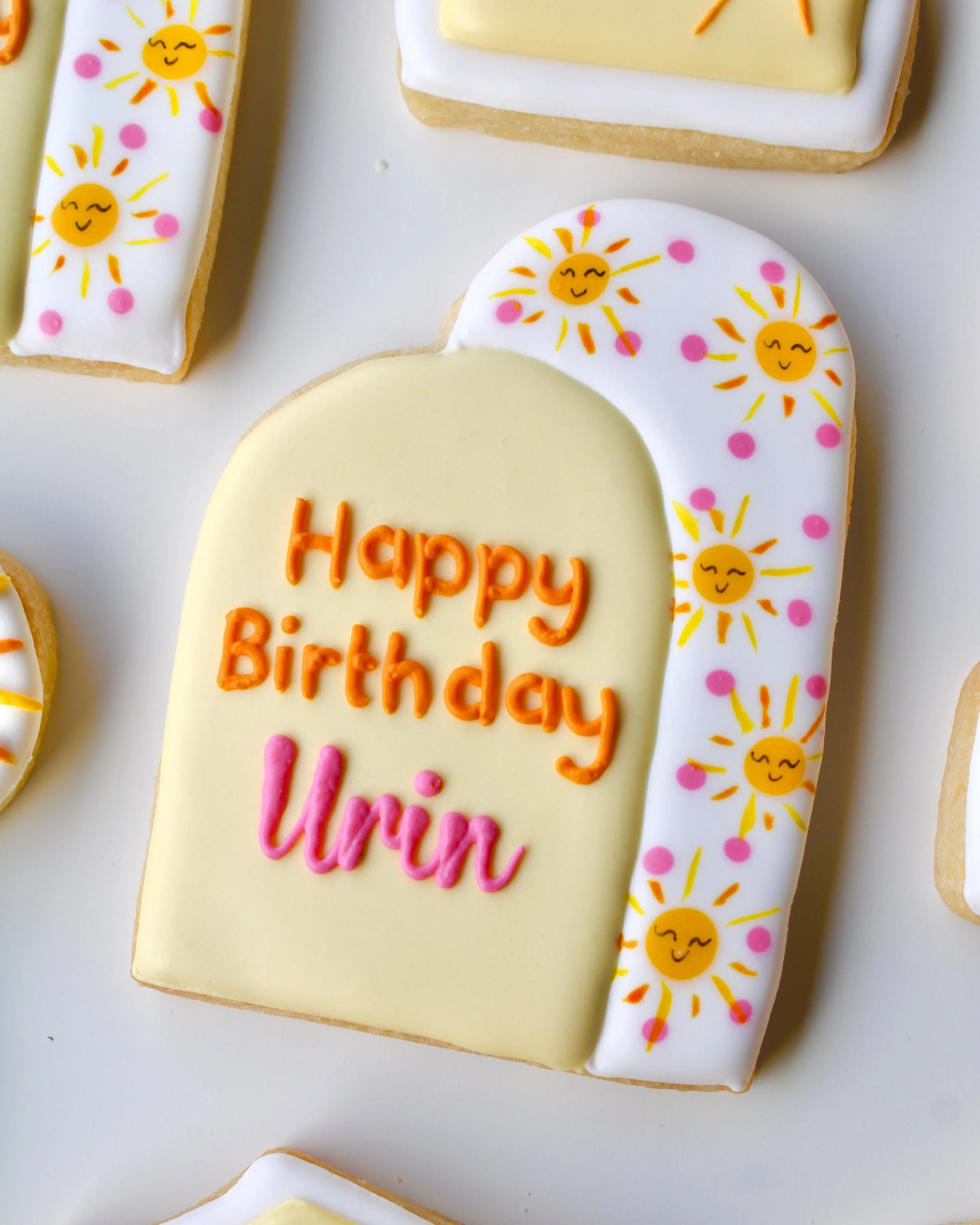 Close-up of a double arch design cookie with hand-painted suns and the words “Happy Birthday Urin” written in bright orange and pink icing.