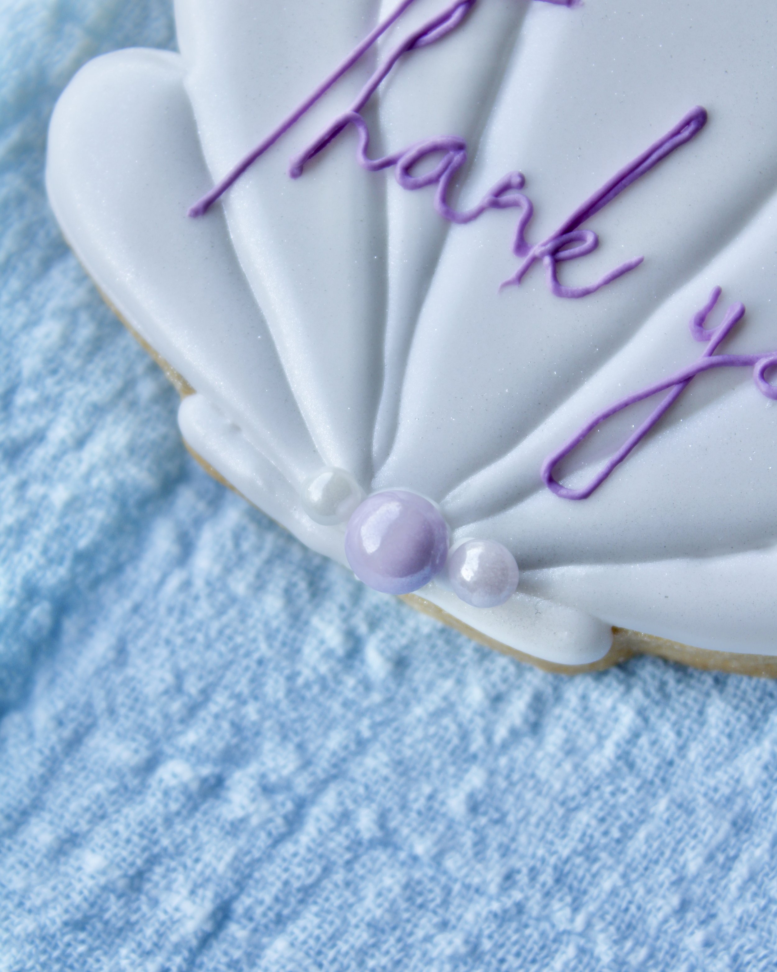 Close up of white seashell cookie with “thank you” written in purple cursive icing, placed on blue linen napkin.